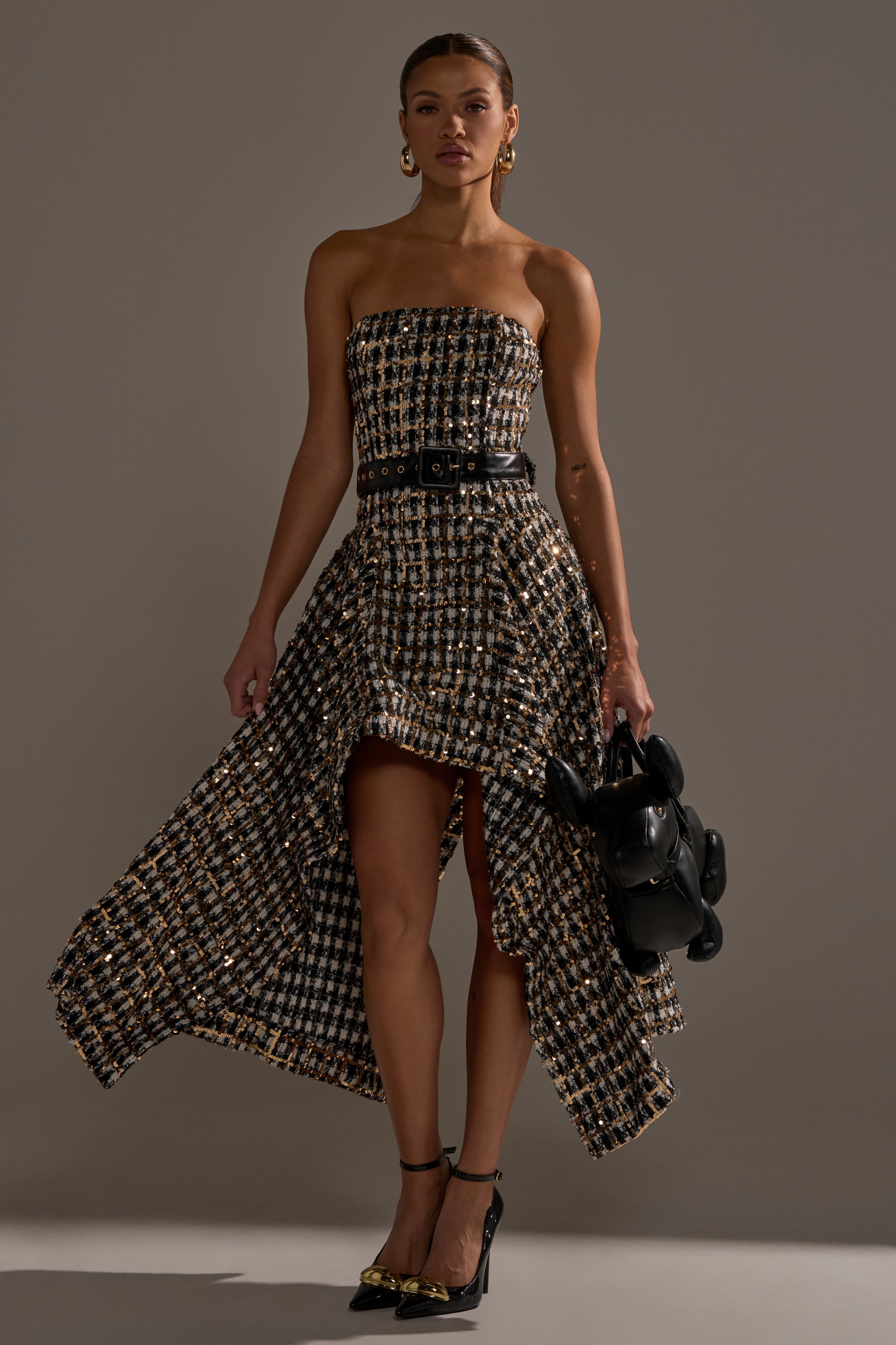 A woman wears the PURE ROYALTY BROCADE HIGH LOW TWEED DRESS WITH SEQUINS, cinched with a black belt. She pairs it with black heels featuring gold accents, gold hoop earrings, and a small black dog-shaped bag against a gray background.