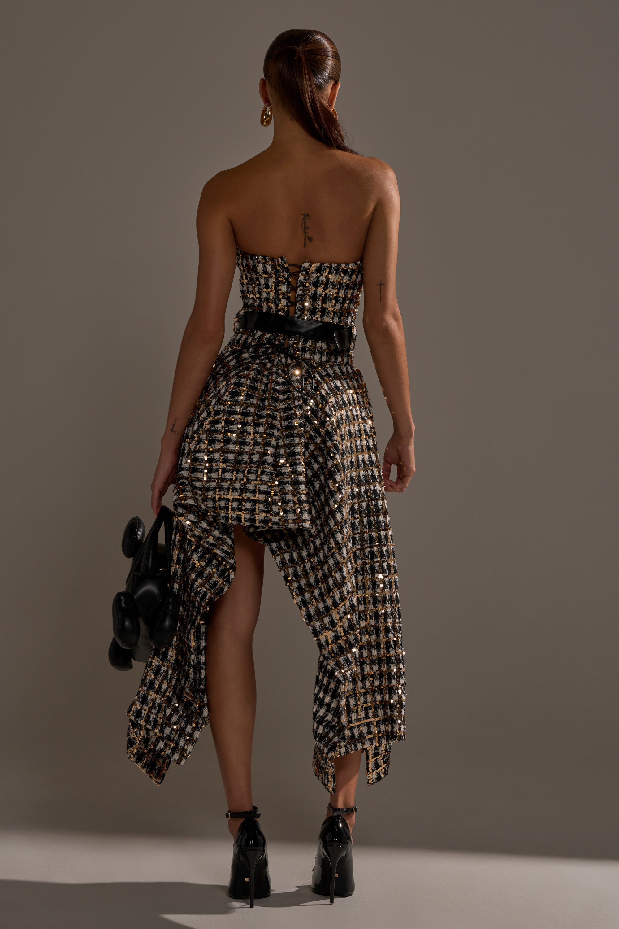 A woman with straight brown hair in a ponytail wears the PURE ROYALTY BROCADE HIGH LOW TWEED DRESS WITH SEQUINS in gold and black plaid, holding a black handbag against a plain gray background.