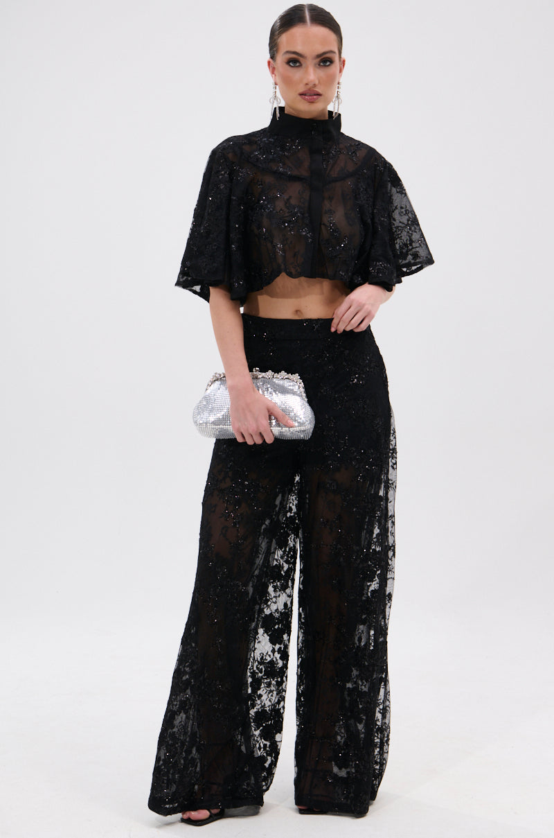A woman stands against a white background wearing the QUEEN B LACE TROUSER and a matching black sheer floral mesh, sequined crop top with wide sleeves. She holds a silver clutch and her hair is pulled back.