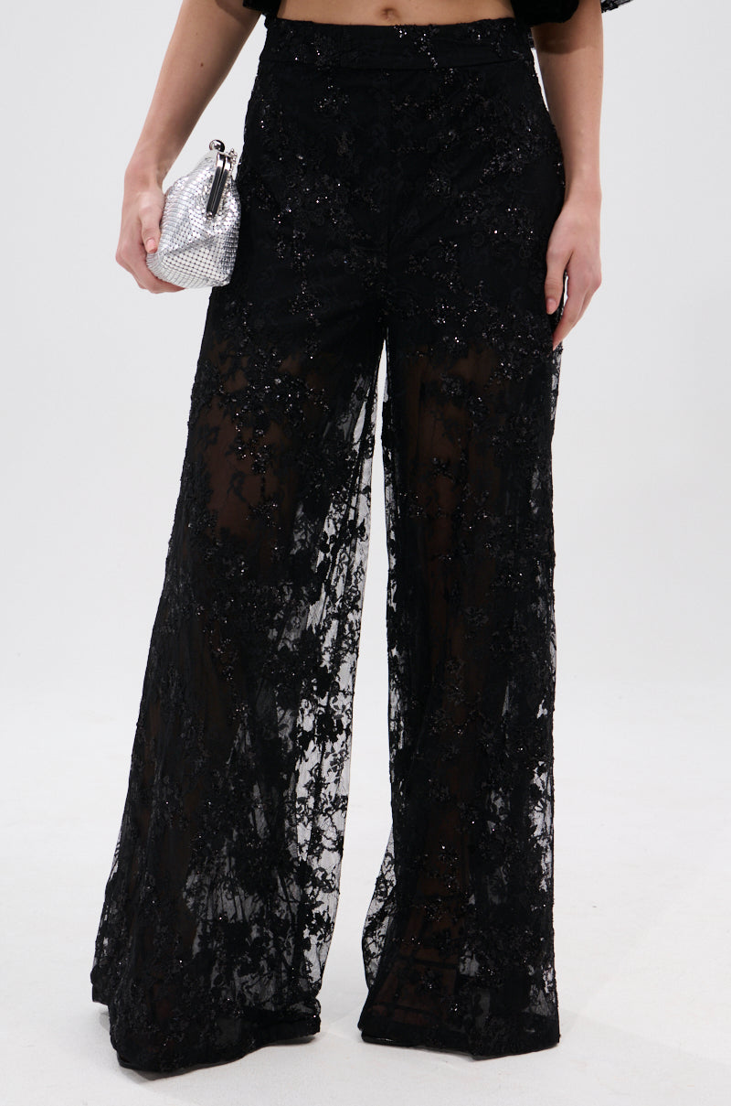 Wearing the QUEEN B LACE TROUSER—high-rise, wide-leg lace trousers with sequins and sheer floral mesh—a person holds a silver textured clutch against a plain, light background.