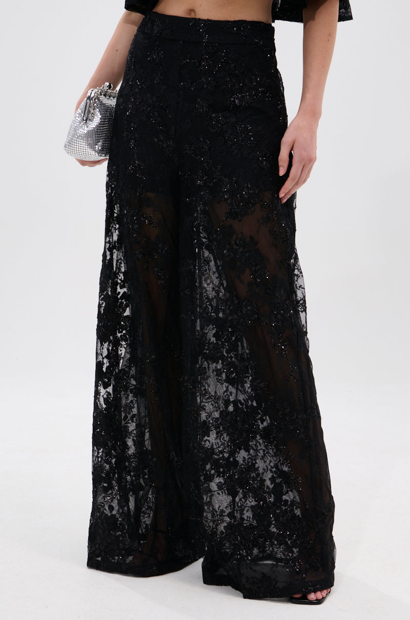 Wearing the QUEEN B LACE TROUSER—high-rise, wide-leg lace pants in sheer floral mesh with sequin details—a person holds a small silver purse against a plain white background.