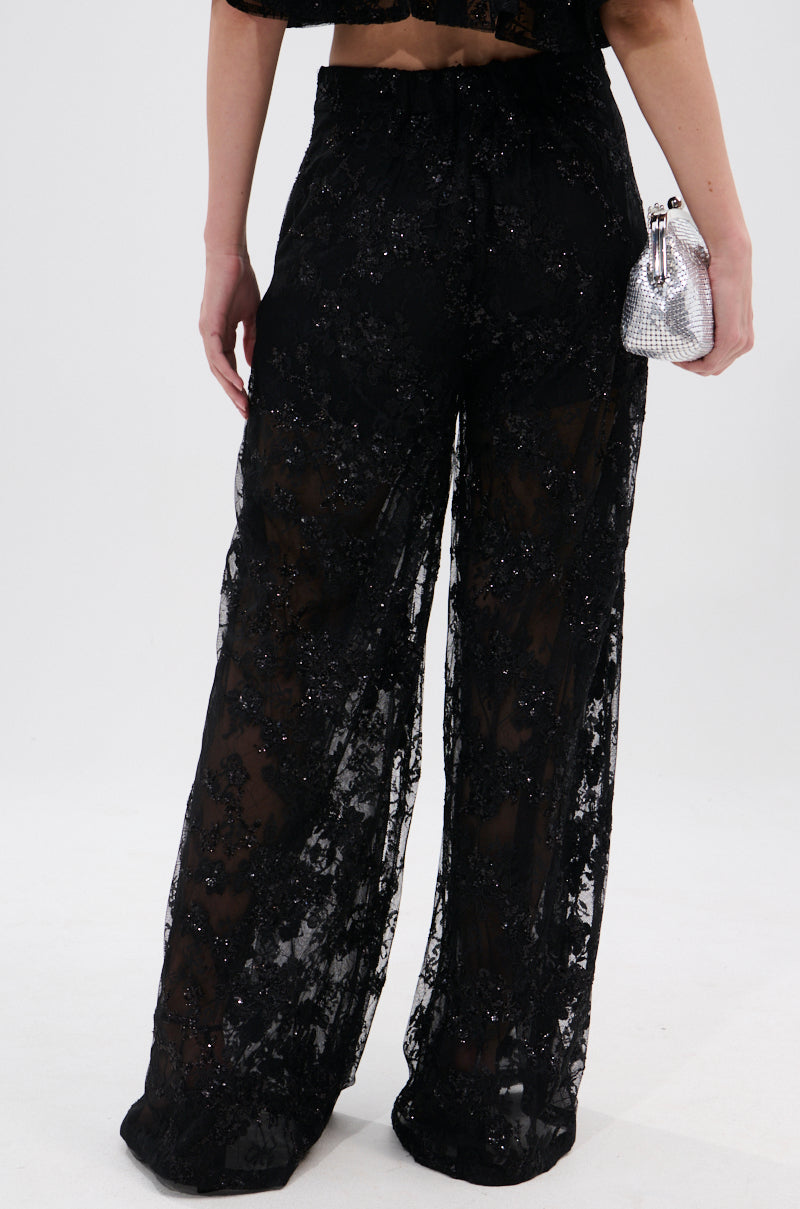 A person faces away, wearing QUEEN B LACE TROUSER high-rise wide-leg sheer black sequin pants with a matching top, holding a small silver textured handbag in their right hand against a plain white background.