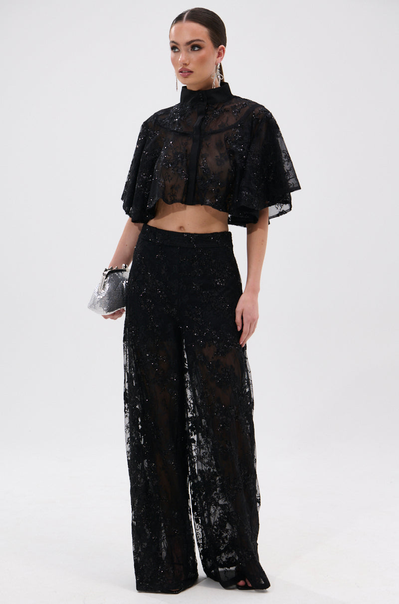 A woman wears QUEEN B LACE TROUSER, paired with a sheer black floral mesh sequined crop top featuring a high neck and cape sleeves. She accessorizes with statement earrings and a silver clutch against a plain white background.