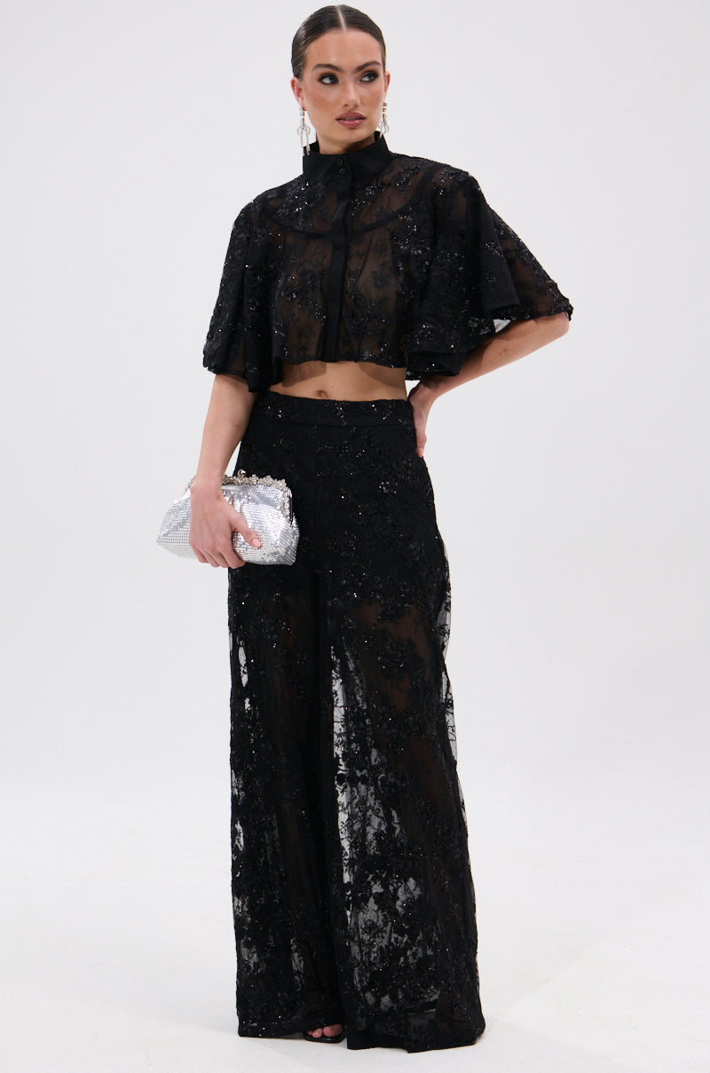 A woman stands against a plain background wearing the QUEEN B LACE TROUSER, a sheer floral mesh two-piece with high-rise wide-leg pants and a cropped top. She holds a silver clutch, her hair pulled back to highlight her statement earrings.