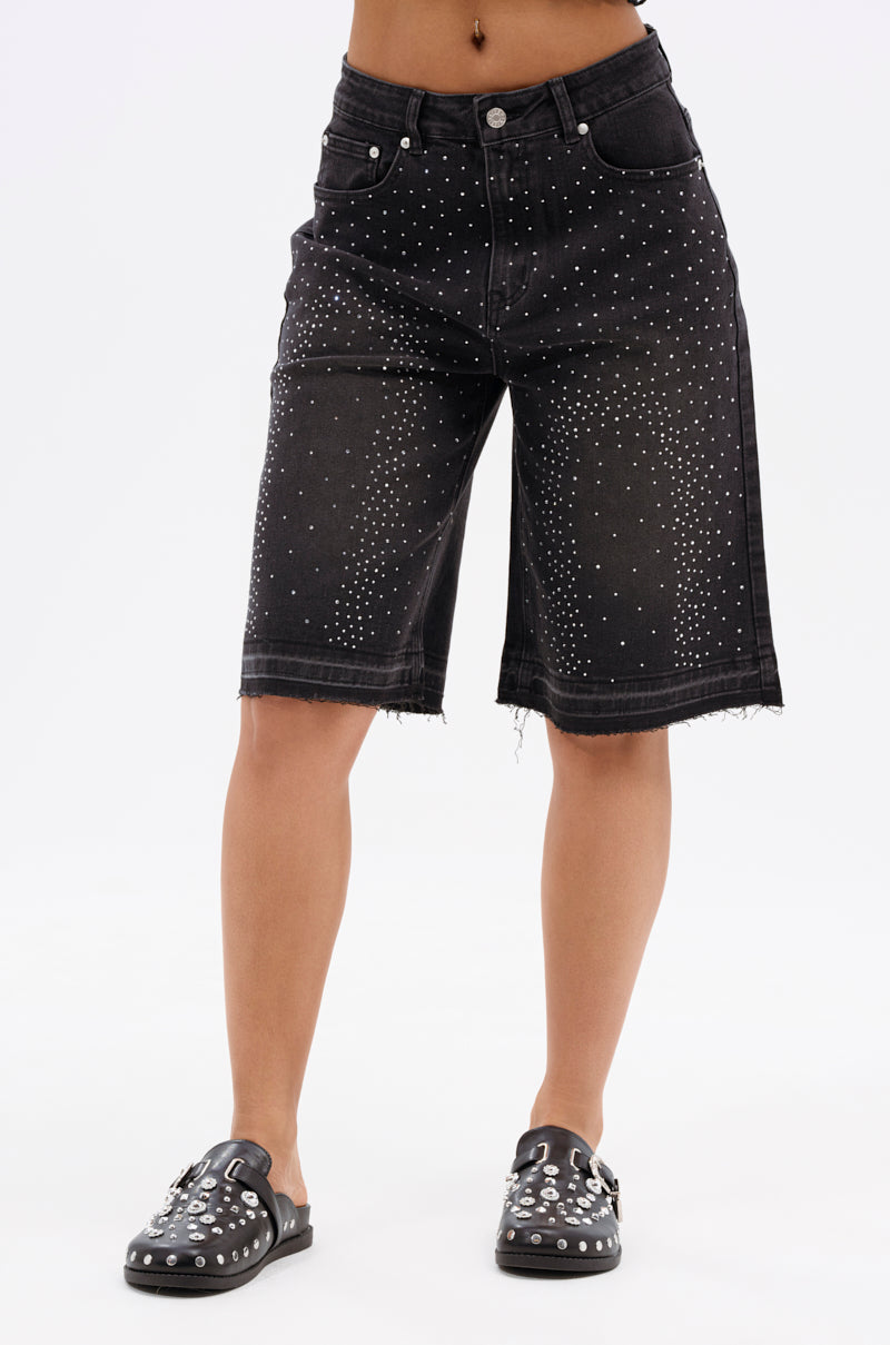A person wearing the STARS SHINING RHINESTONE BERMUDA SHORT—black, mid-rise stretchy shorts with silver studs and a frayed hem—plus black slip-on shoes with silver studs, stands against a plain white background.