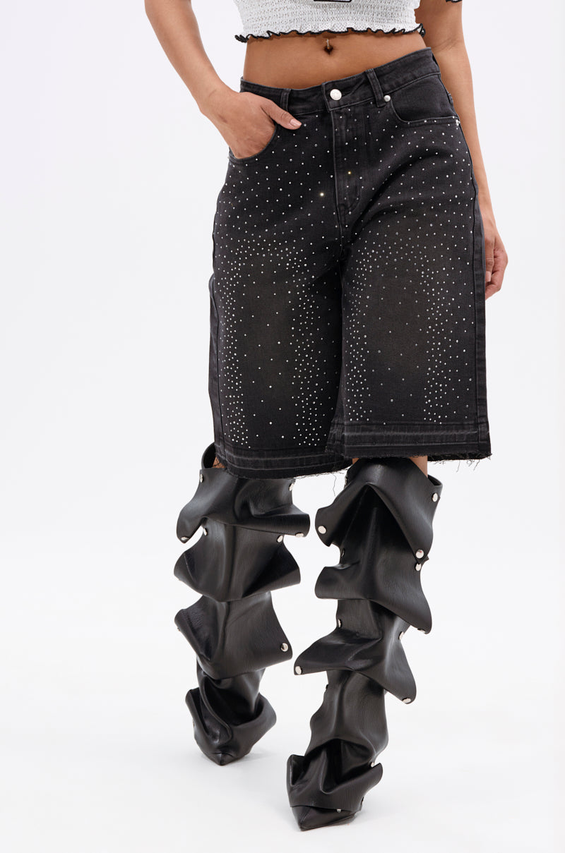 A person wearing STARS SHINING RHINESTONE BERMUDA SHORTS paired with unique black sculptural boots featuring layered, ruffled sections. Their hand rests in a pocket, and only the torso and legs are visible.