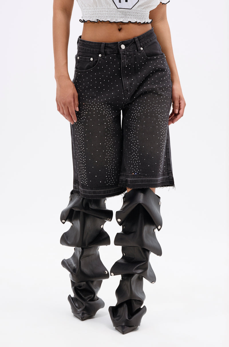 A person wearing a white crop top, STARS SHINING RHINESTONE BERMUDA SHORT, and unique black ruffled thigh-high boots with metal rivets, standing against a plain white background.