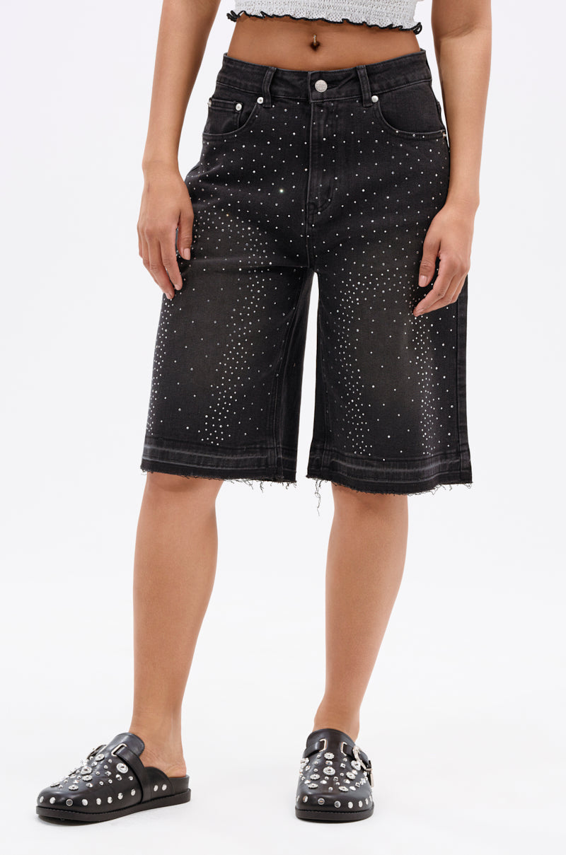 A person wearing the STARS SHINING RHINESTONE BERMUDA SHORT—black, mid-rise, stretchy denim shorts with silver rhinestones and a frayed hem—and matching black slip-on shoes with silver studs, standing against a plain white background.