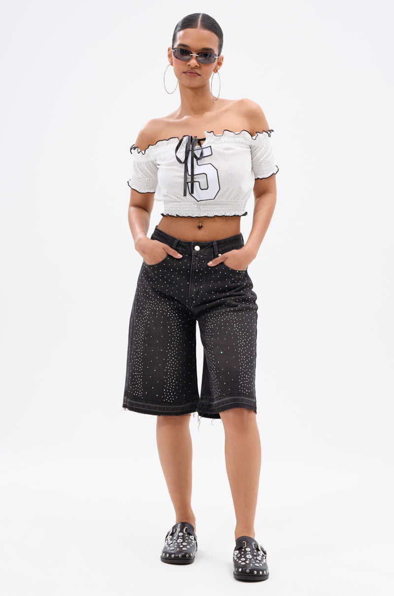 A woman completes her streetwear look in sunglasses, hoop earrings, a white off-shoulder "6" crop top, studded black slip-ons, and the STARS SHINING RHINESTONE BERMUDA SHORT with hands in her pockets against a plain background.