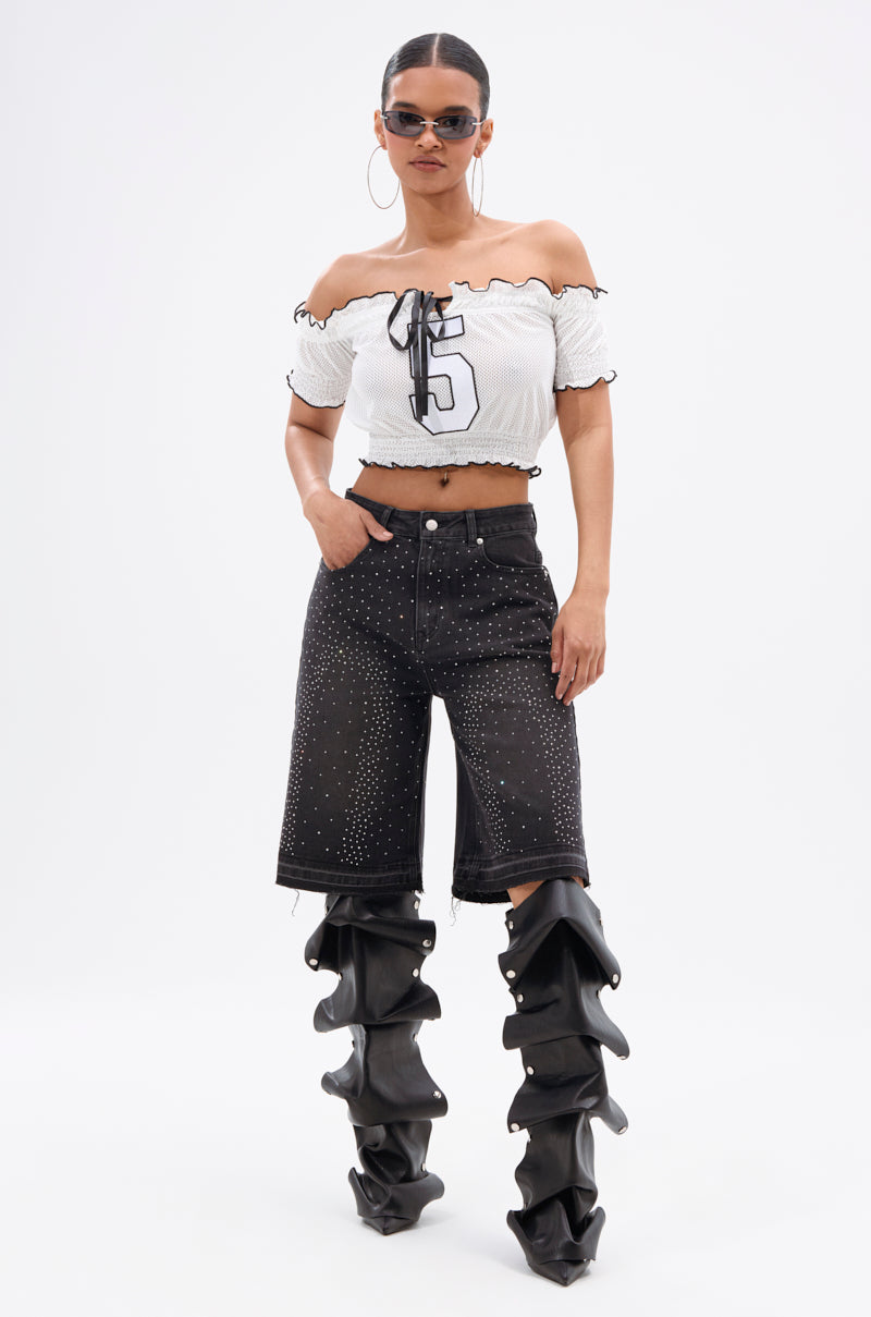 A woman wears the STARS SHINING RHINESTONE BERMUDA SHORT with a white off-shoulder "5" crop top, oversized black ruched boots, large hoop earrings, and sunglasses, posing against a plain white background.