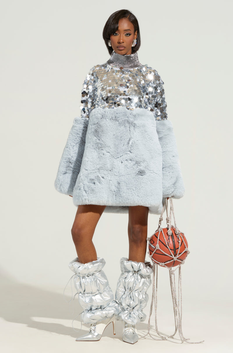 A model poses in a stylish outfit comprising a shiny, oversized, silver-sequined top and a light gray faux fur skirt. Her OUTTA THIS WORLD PUFFER LEGWARMERS coordinate perfectly with the unique orange spherical handbag she holds, which is adorned with long tassels.