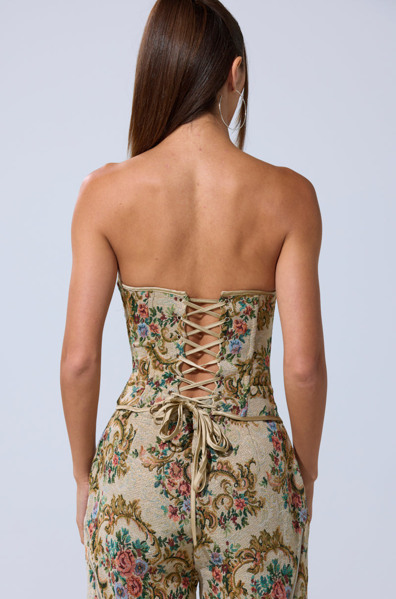 A woman with long brown hair is seen from behind, wearing the SHINE AWAY BROCADE CORSET— a strapless floral corset with lace-up detail and delicate pattern—paired with matching pants, set against a plain light background.