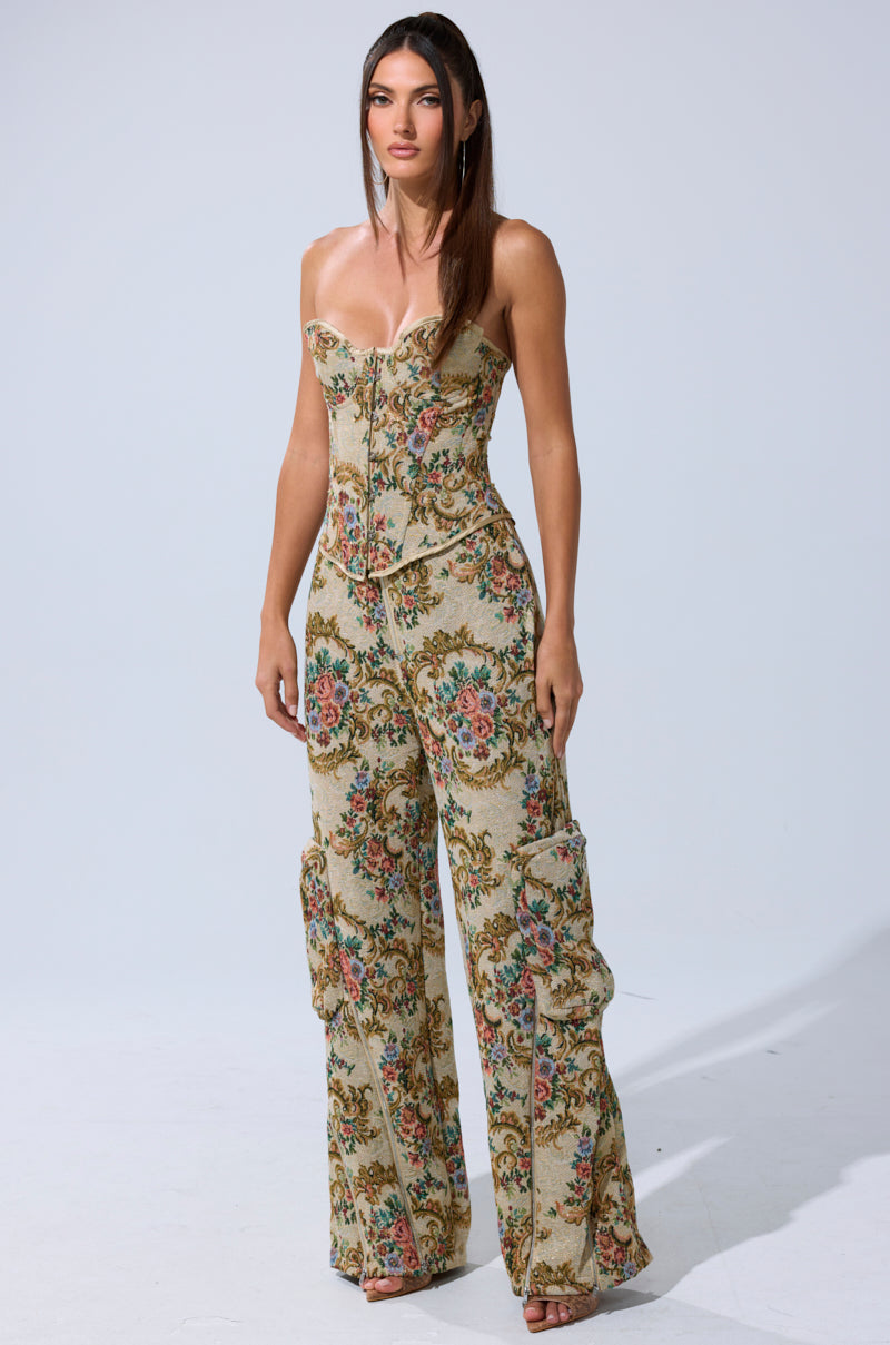 A woman stands facing forward against a plain light background, wearing the SHINE AWAY BROCADE CORSET and wide-leg pants with a baroque floral tapestry pattern in beige, green, pink, and blue tones.