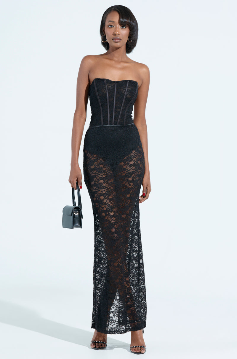 A woman stands confidently in the TRENDSETTER LACE MAXI DRESS, boasting a strapless design and stylish lace-up back. The structured bodice and sheer skirt pair elegantly with her high heels and small black handbag against the plain, light background.