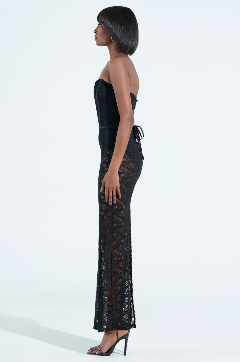 A woman poses in profile against a light background, showcasing the TRENDSETTER LACE MAXI DRESS. The strapless design highlights her elegant form, complemented by high heels. Her sleek bob hairstyle enhances her sophistication, and a subtle lace-up back closure completes the look.