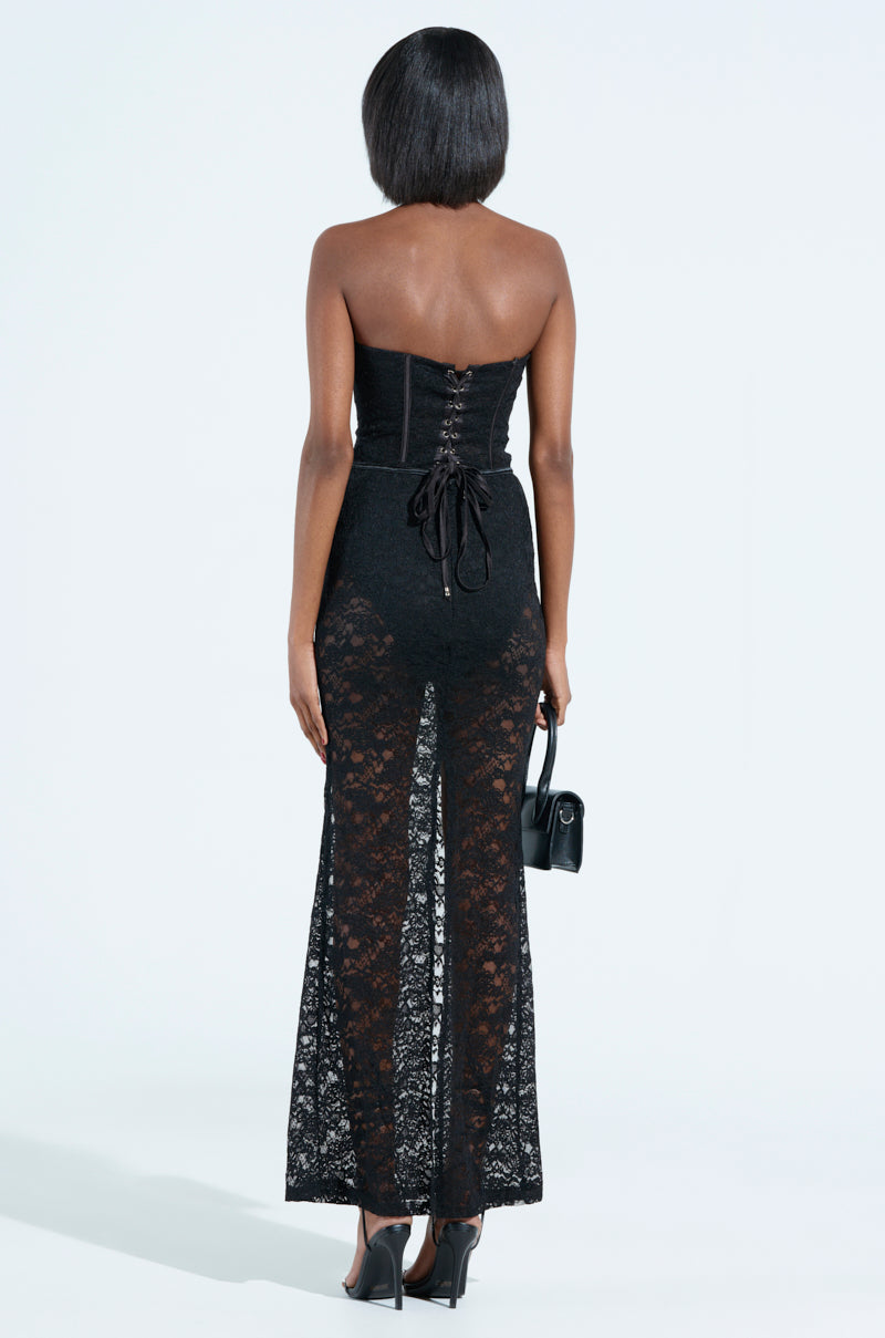 A person in a strapless silhouette wears the TRENDSETTER LACE MAXI DRESS against a white background. With a black lace-up back, they hold a small black purse and don sleek heels, exuding effortless elegance and style.