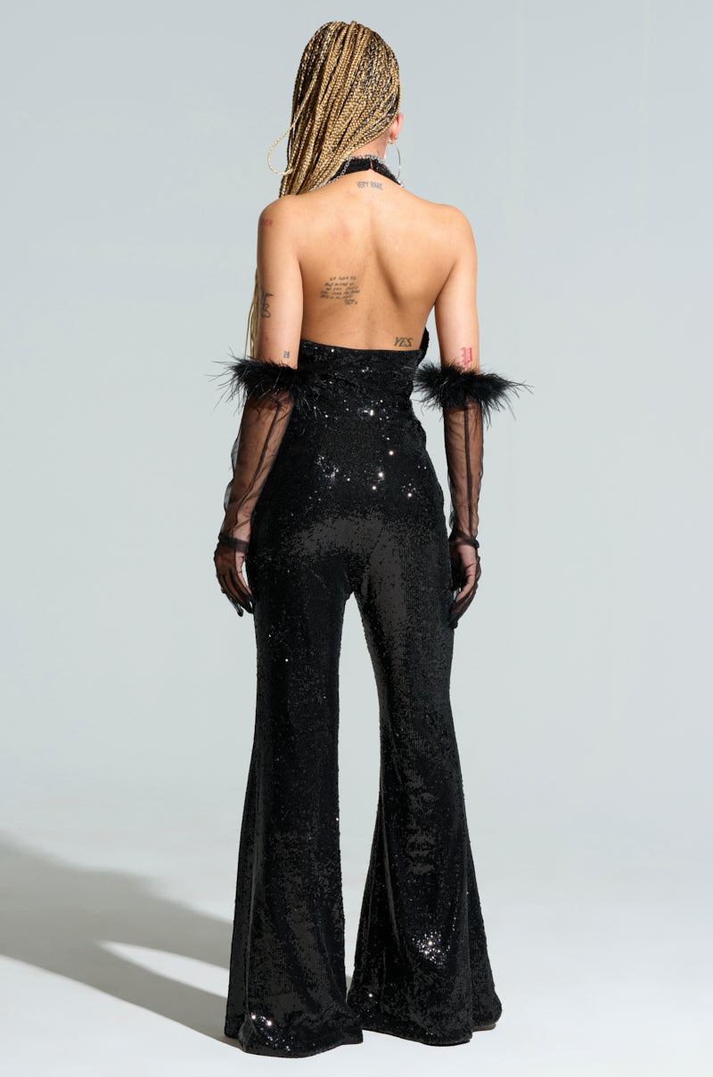 A person with braided hair is wearing the WE WILL ROCK YOU SEQUIN MESH JUMPSUIT, paired with feathered gloves. They stand with their back to the camera, highlighting tattoos on their upper back and shoulders, while the outfit elegantly accentuates a subtle flare leg silhouette.