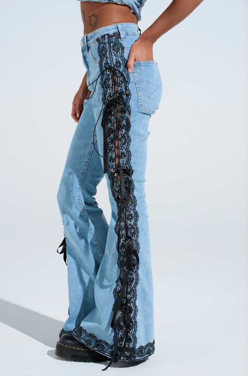 The image features a person in TICKED AND TIED LACE AND BOW DETAIL DENIM PANTS with black lace-up panels, flared leg silhouette, and black lace trim. They complete the look with black shoes, a cropped top, and tattoos on their midriff.