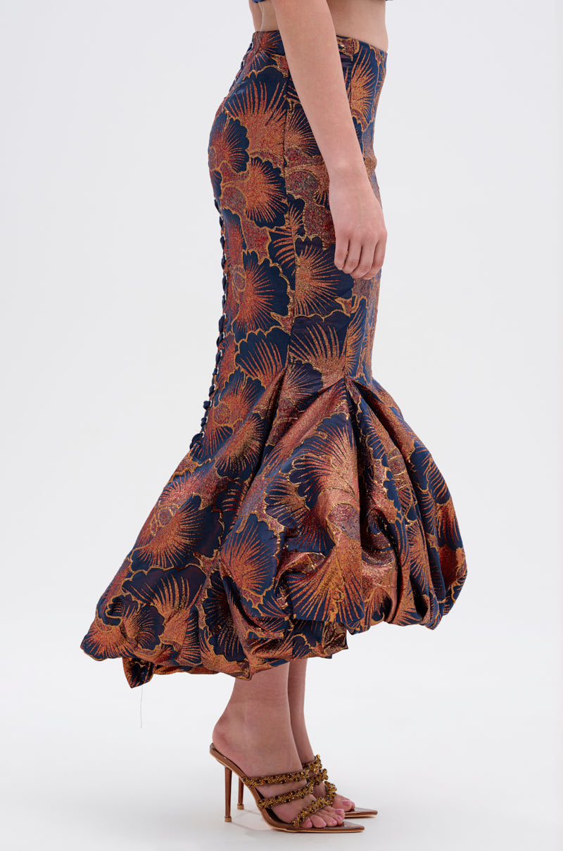 A woman stands sideways wearing the LADY LUX BROCADE BUBBLE HEM MIDI SKIRT, a fitted high-rise design with a bold copper and navy metallic floral pattern. The adjustable lace-up back and strappy heeled sandals complete her refined look.