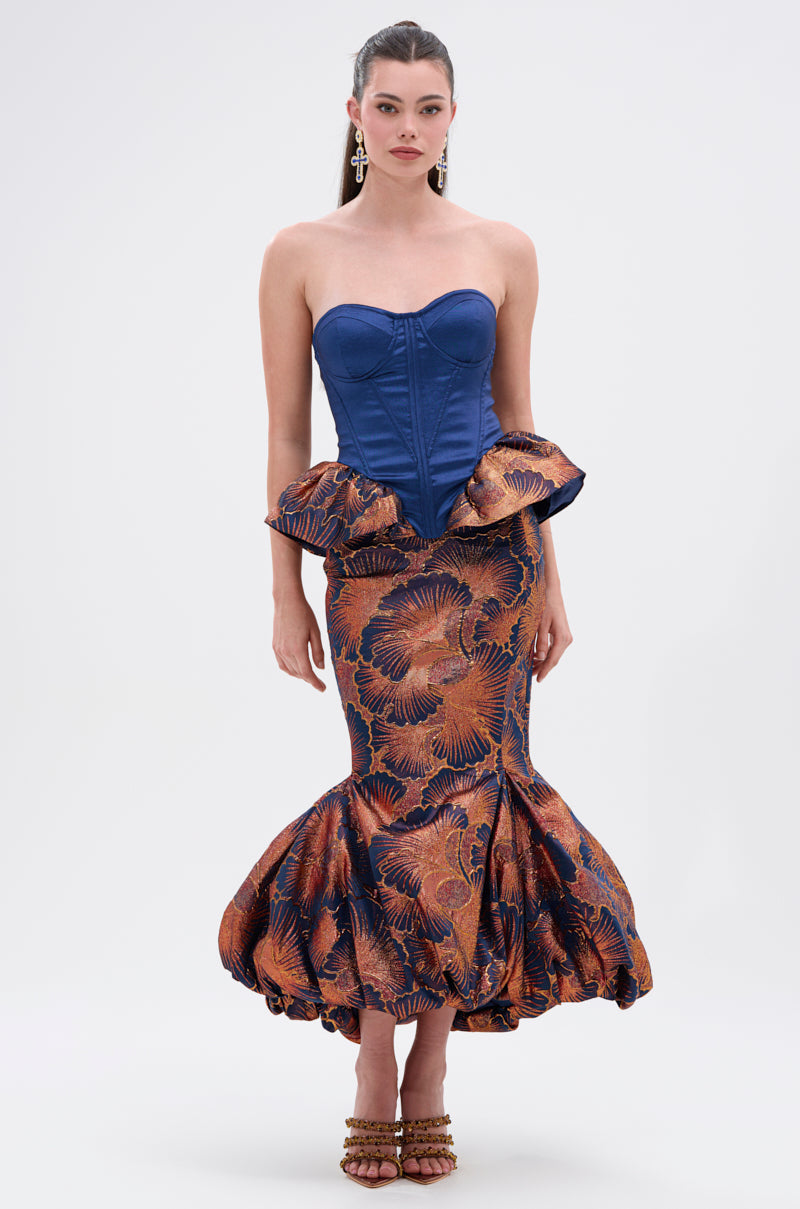 A woman wears statement earrings and the LADY LUX BROCADE BUBBLE HEM MIDI SKIRT, a high-rise silhouette with dramatic bubble hem, adorned with bold orange and blue florals, paired with a strapless blue corset top featuring an adjustable lace-up back.
