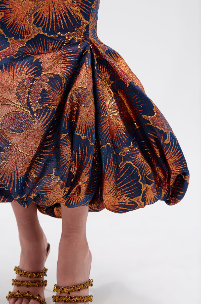 A close-up of a person wearing the LADY LUX BROCADE BUBBLE HEM MIDI SKIRT with metallic gold and orange dark floral brocade, a dramatic bubble hem, and strappy embellished heels.