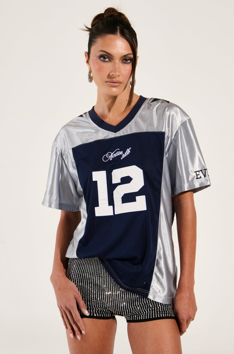 A person exudes sporty chic in the SLICK JERSEY, a sleek silver and navy blue garment featuring the number 12. Their dark hair is pulled back, perfectly complementing black and white colorblock patterned shorts. The jersey proudly displays "Autumn Wolf" and "EVE" in bold letters, embodying a confident athletic style.