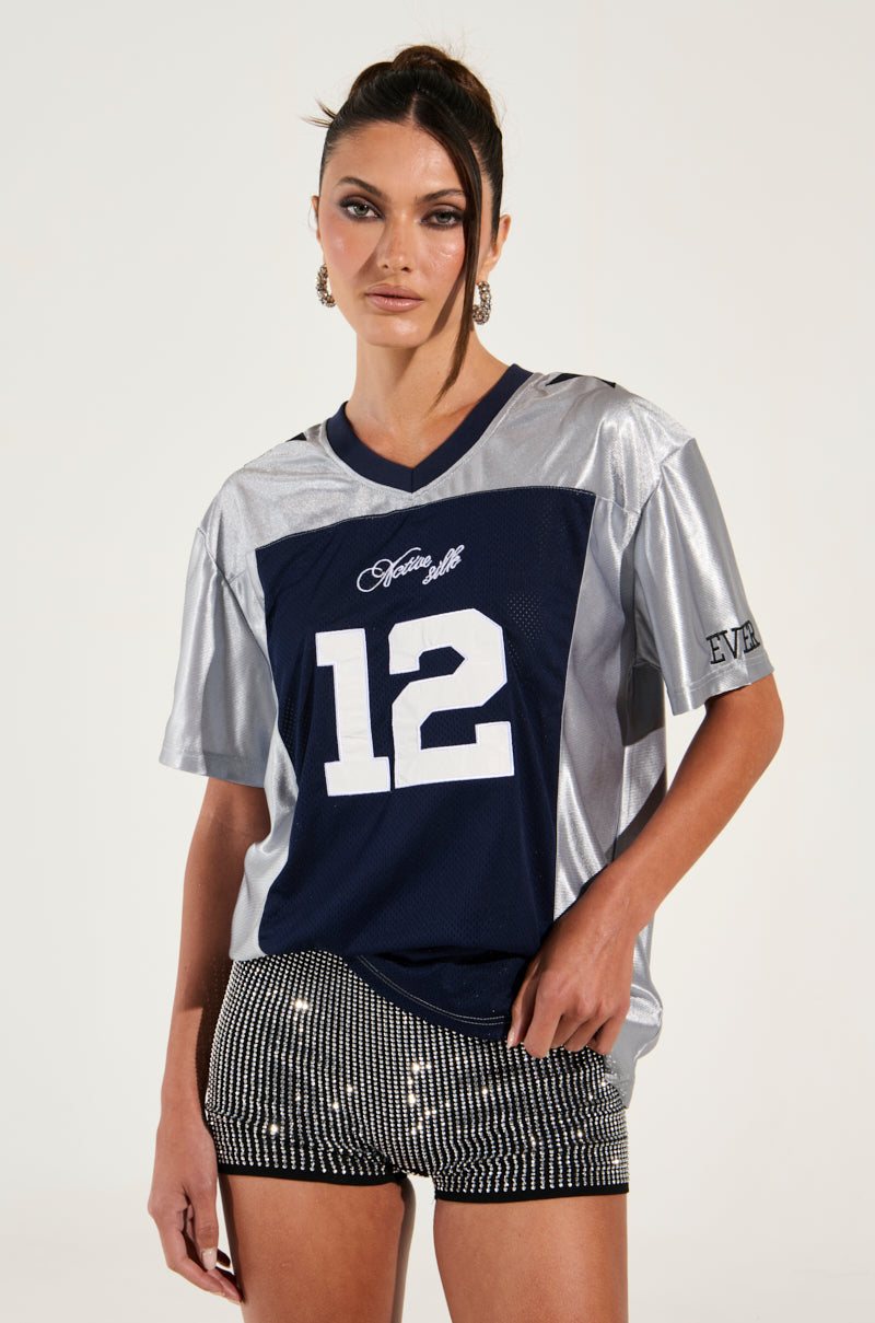 A woman stands confidently against a neutral background, epitomizing sporty chic. She is dressed in the SLICK JERSEY, featuring a blue and silver colorblock pattern with the number 12 on it, paired with metallic shorts adorned with sparkling details. Her hair is tied back, and she wears statement earrings, showcasing her sleek style.