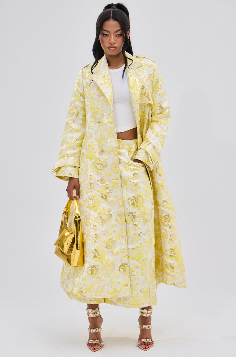 A woman stands against a plain background wearing the NO EXPECTATIONS BROCADE TRENCH—a long, yellow coat with metallic floral motifs—over a white crop top and skirt, accessorized with a gold handbag and metallic strappy heels.