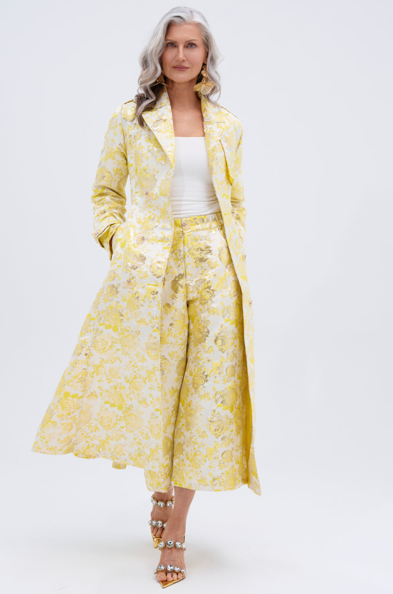 A confident older woman with long gray hair wears the NO EXPECTATIONS BROCADE TRENCH and matching culotte pants, styled with a white top and jeweled heels, standing against a plain white background.