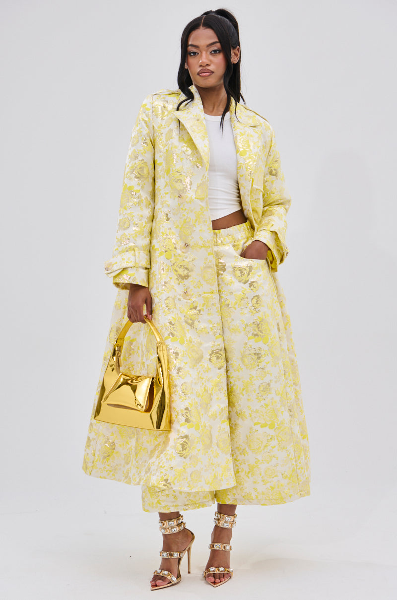 A woman models the NO EXPECTATIONS BROCADE TRENCH in yellow with metallic floral patterns, styled with matching wide-leg pants, a white crop top, gold handbag, and metallic strappy high heels.