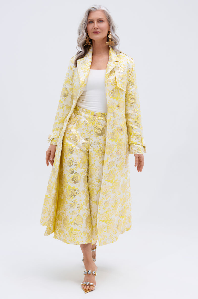 A woman with long gray hair confidently poses against a white background in the NO EXPECTATIONS BROCADE TRENCH—featuring metallic floral motifs and a double-breasted front—styled with matching wide-leg pants and open-toe heels.