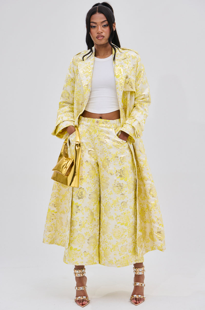 A woman wears the NO EXPECTATIONS BROCADE TRENCH—long, yellow with metallic floral motifs and double-breasted front—paired with matching wide-leg pants, a white crop top, gold handbag, and strappy heels against a white background.