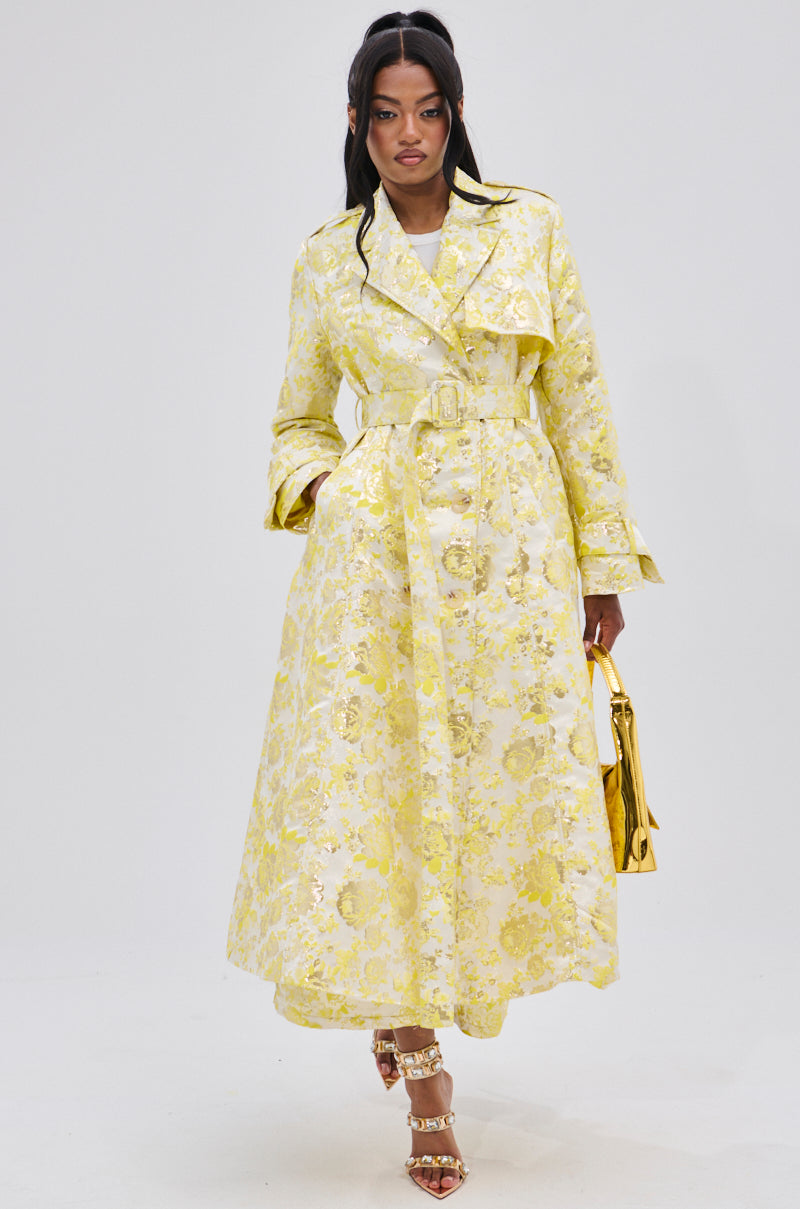 A woman confidently poses against a plain white background wearing the NO EXPECTATIONS BROCADE TRENCH with metallic floral motifs and a double-breasted front, accessorized with a shiny gold handbag and gold strappy heels.