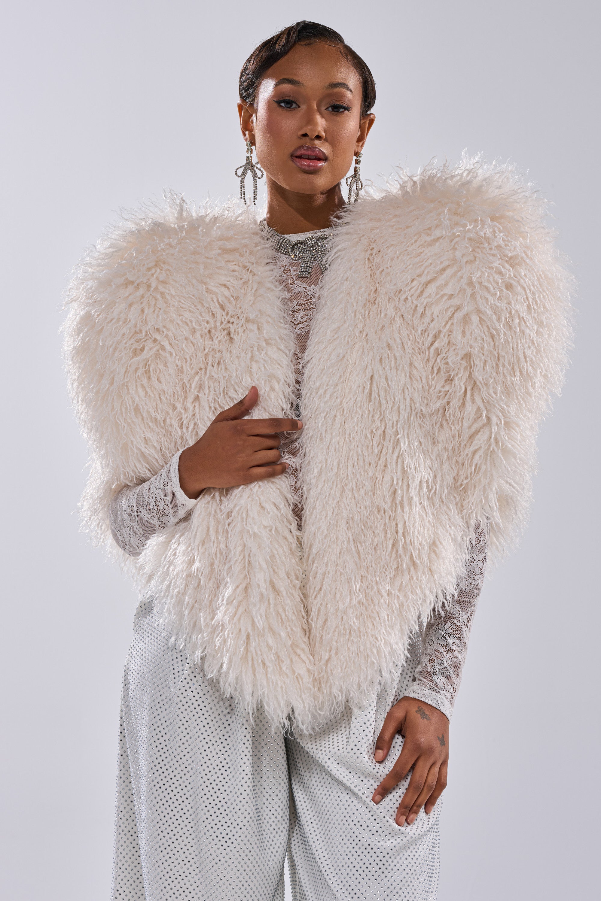 A woman exudes confidence in the STAR CROSSED LOVER FAUX FUR VEST IN IVORY, styled over a lace top and white pants. With statement earrings and chic hair, she poses against a light backdrop—making this vest a true statement piece.