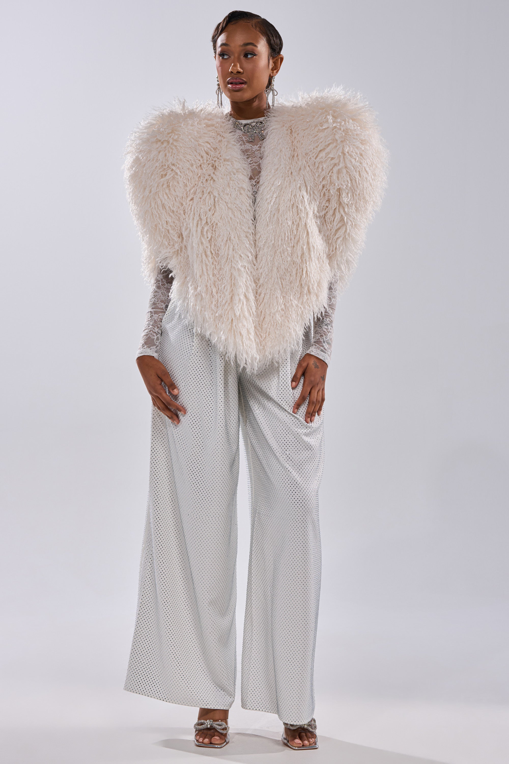 A woman wears the STAR CROSSED LOVER FAUX FUR VEST IN IVORY over silver wide-leg pants and a sheer long-sleeve top, accessorized with earrings, posing confidently against a plain light background.