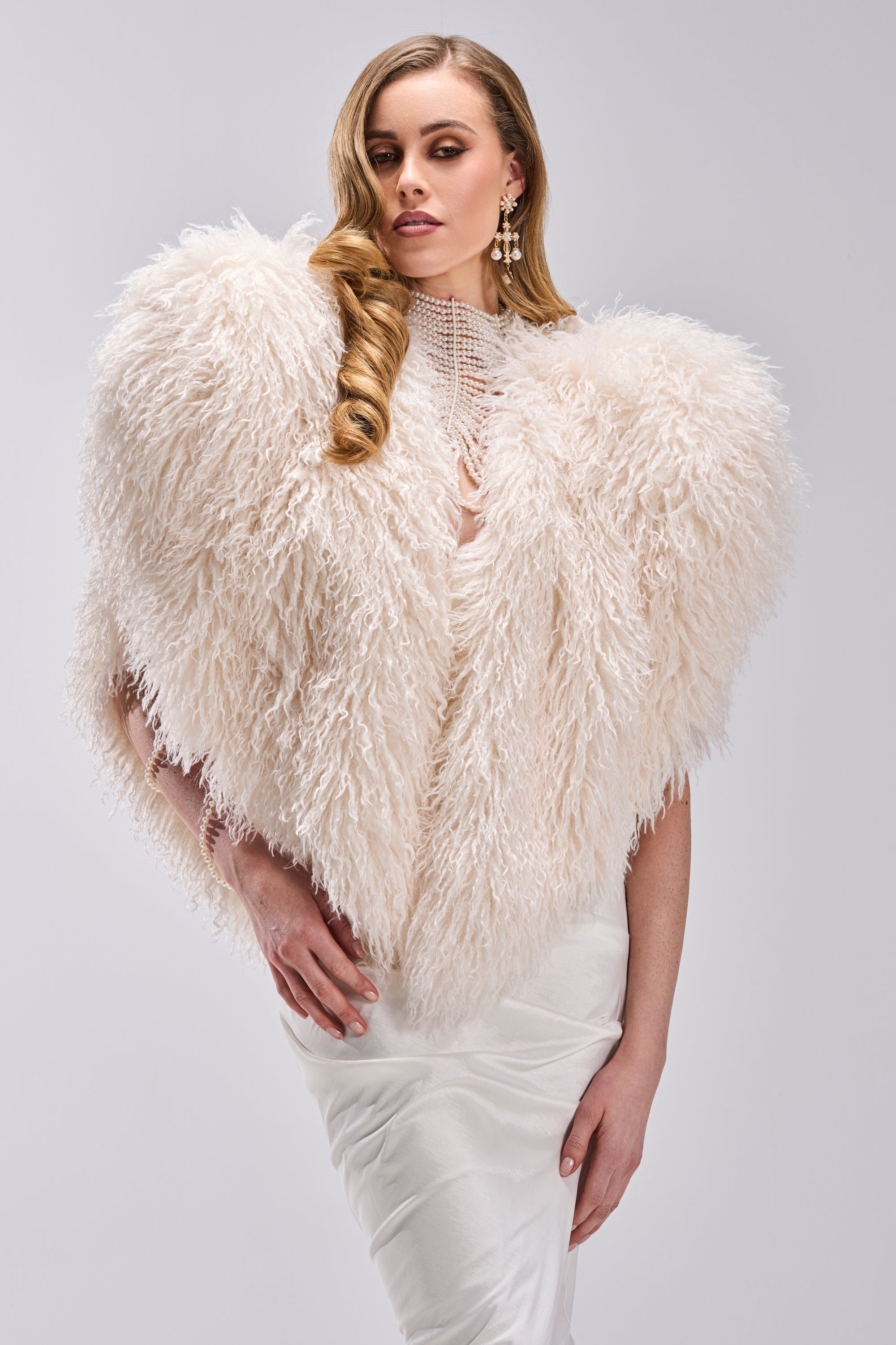A woman models the STAR CROSSED LOVER FAUX FUR VEST IN IVORY over a white satin dress with statement earrings, set against a simple light backdrop.
