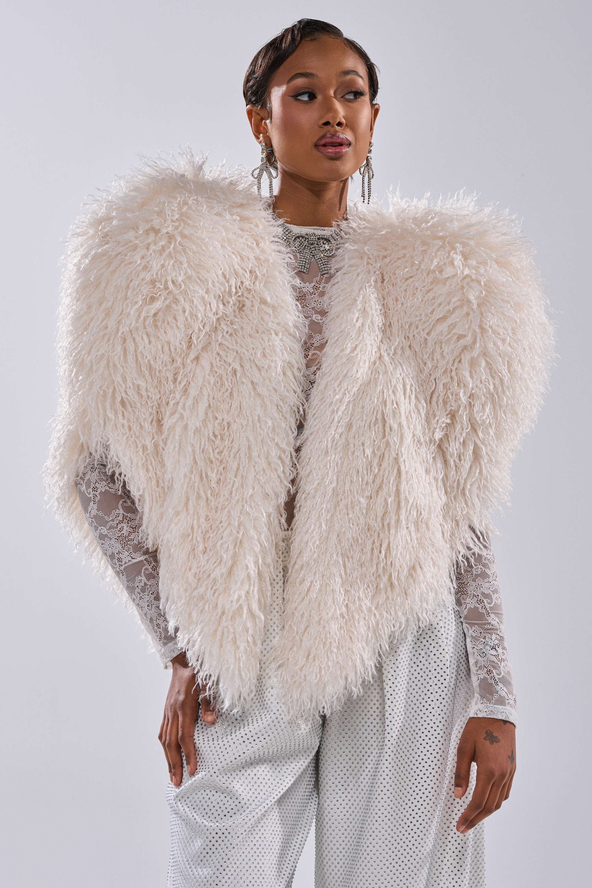 Confidently posing against a plain light background, a person showcases the STAR CROSSED LOVER FAUX FUR VEST IN IVORY over a sheer lace top and white textured pants, accessorized with statement earrings and a necklace for bold style.