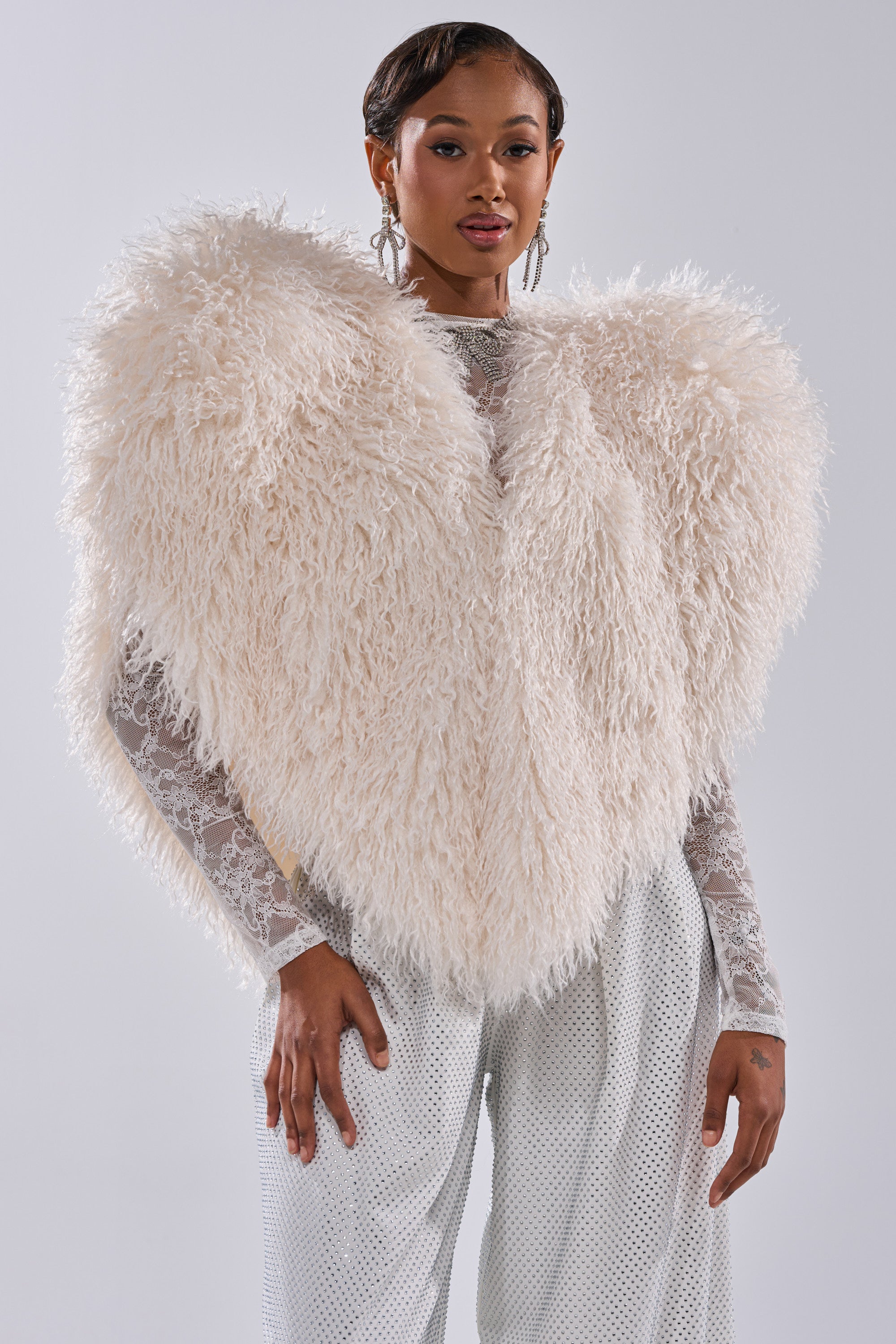 A woman poses confidently against a plain gray background, wearing the STAR CROSSED LOVER FAUX FUR VEST IN IVORY—a striking, wing-like statement piece—layered over a sheer lace top and light dotted pants.