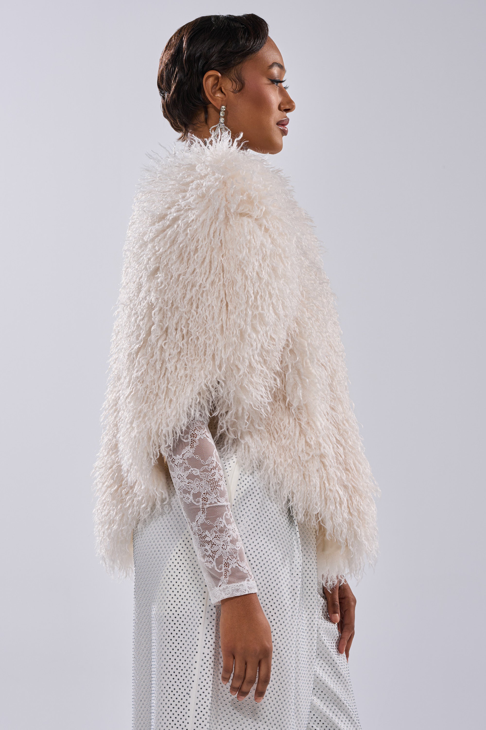 A woman in profile models the STAR CROSSED LOVER FAUX FUR VEST IN IVORY over a white lace top and skirt, posing against a plain light background for a chic, statement look.