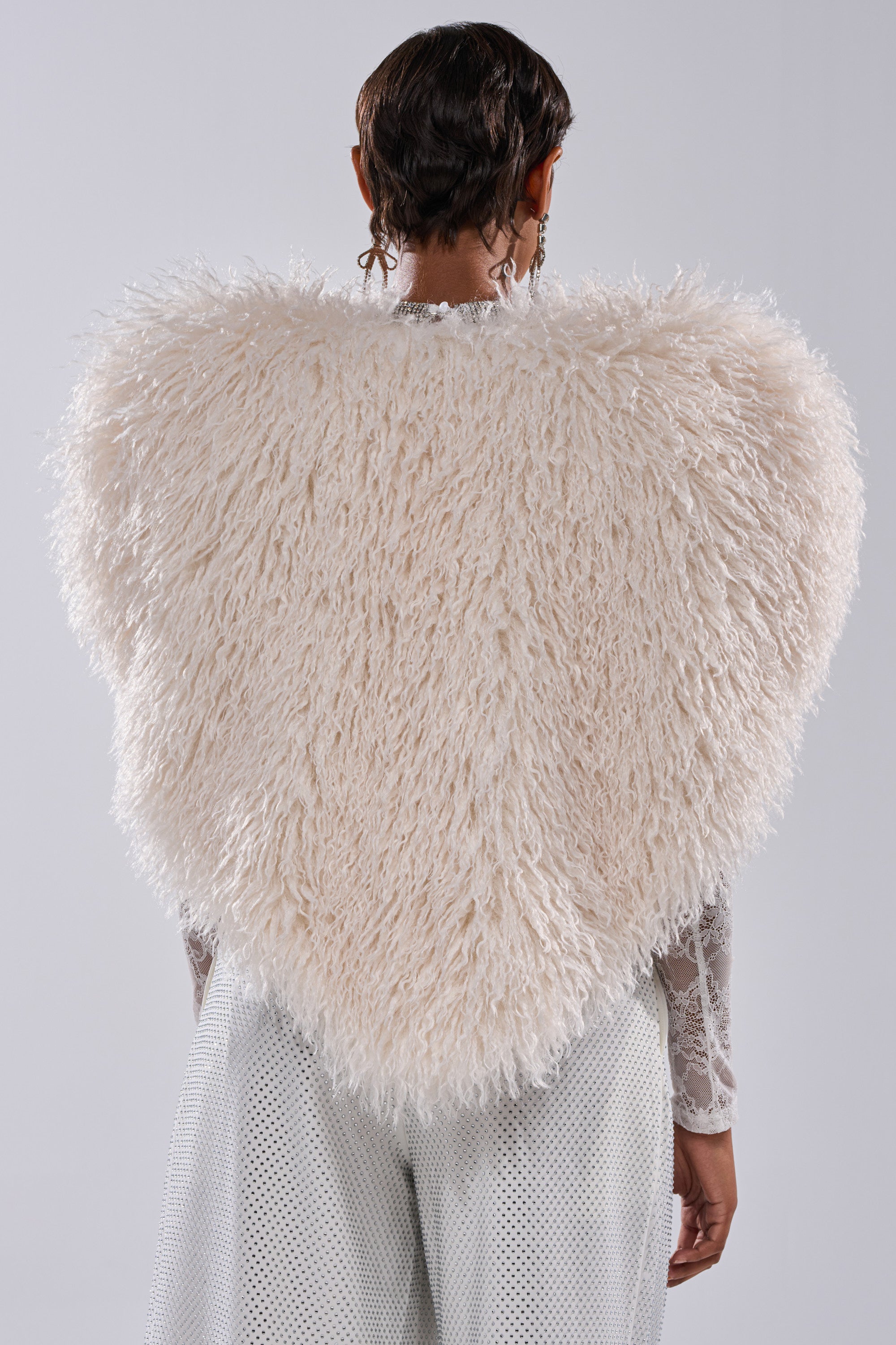 A person seen from behind wears the STAR CROSSED LOVER FAUX FUR VEST IN IVORY—a large, heart-shaped, cream statement piece—over a sheer patterned long-sleeve top and light pants against a plain gray background.