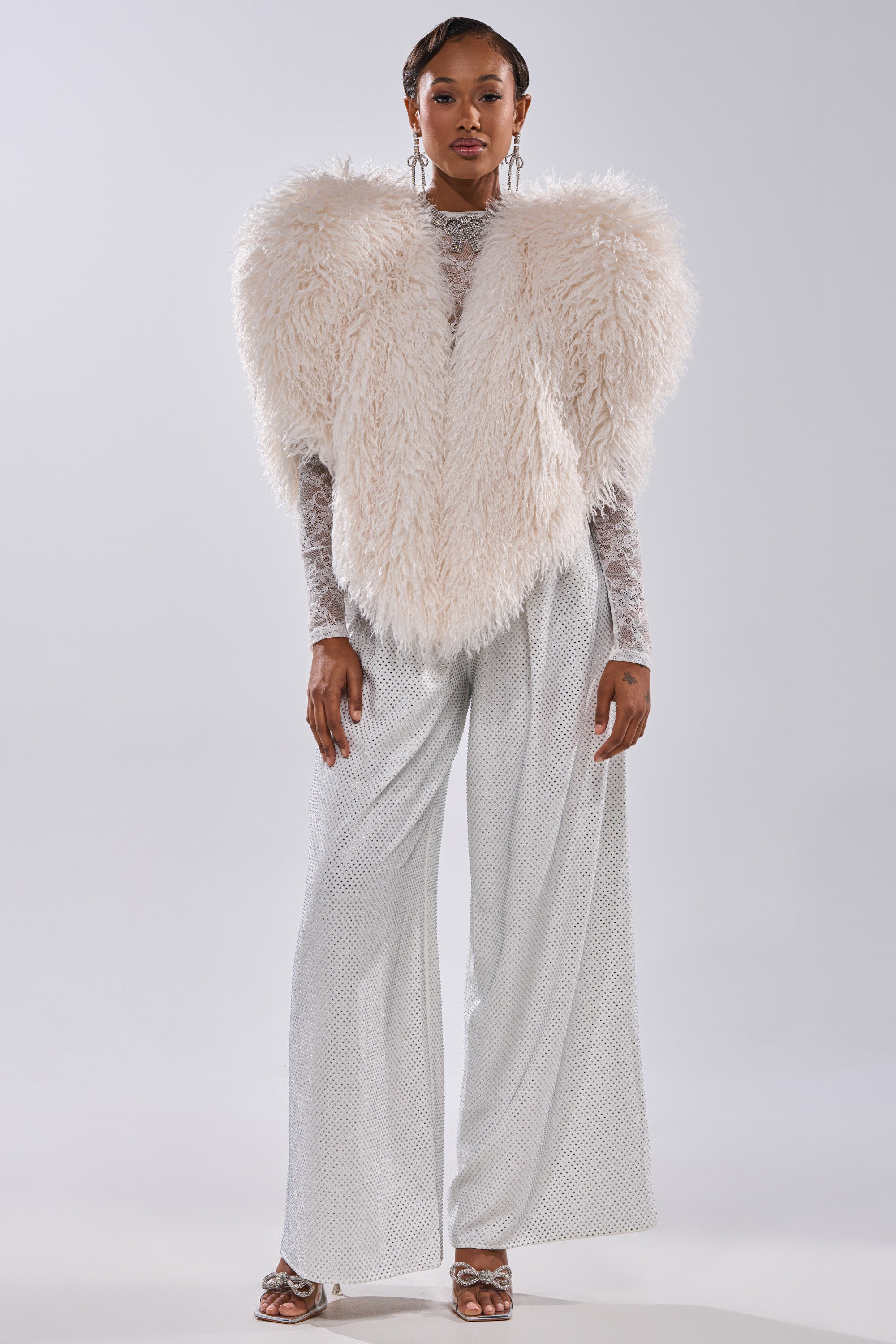 A person faces forward in the STAR CROSSED LOVER FAUX FUR VEST IN IVORY over a sheer long-sleeve top, paired with wide-legged, subtly patterned light gray pants and open-toe heels. Their hair is short and styled.