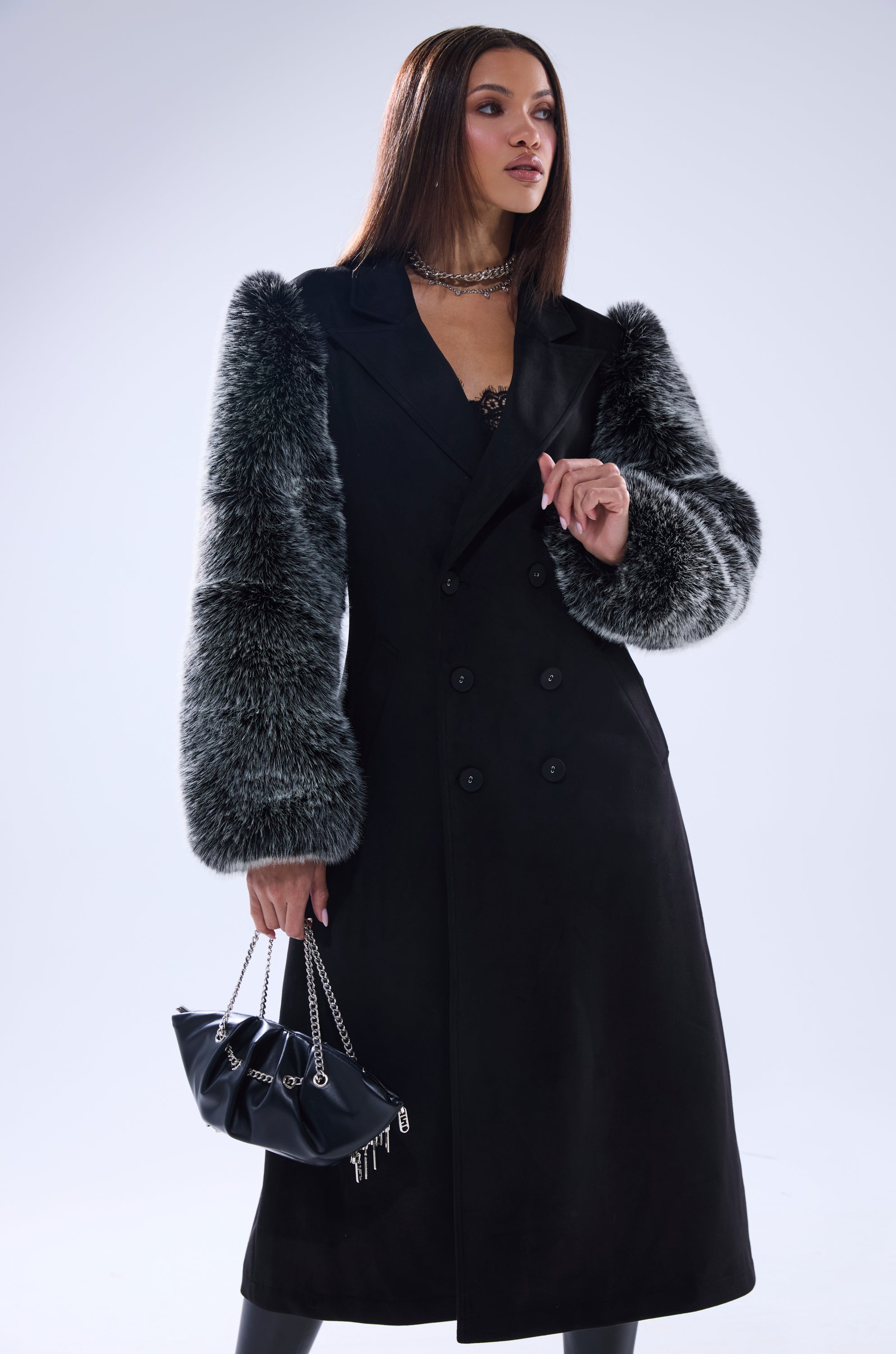 A woman wears the NOLITA FUR SLEEVE TRENCH, a black double-breasted longline coat with gray fur sleeves, paired with a black handbag with silver chains. She has straight brown hair and layered necklaces, standing against a light background.