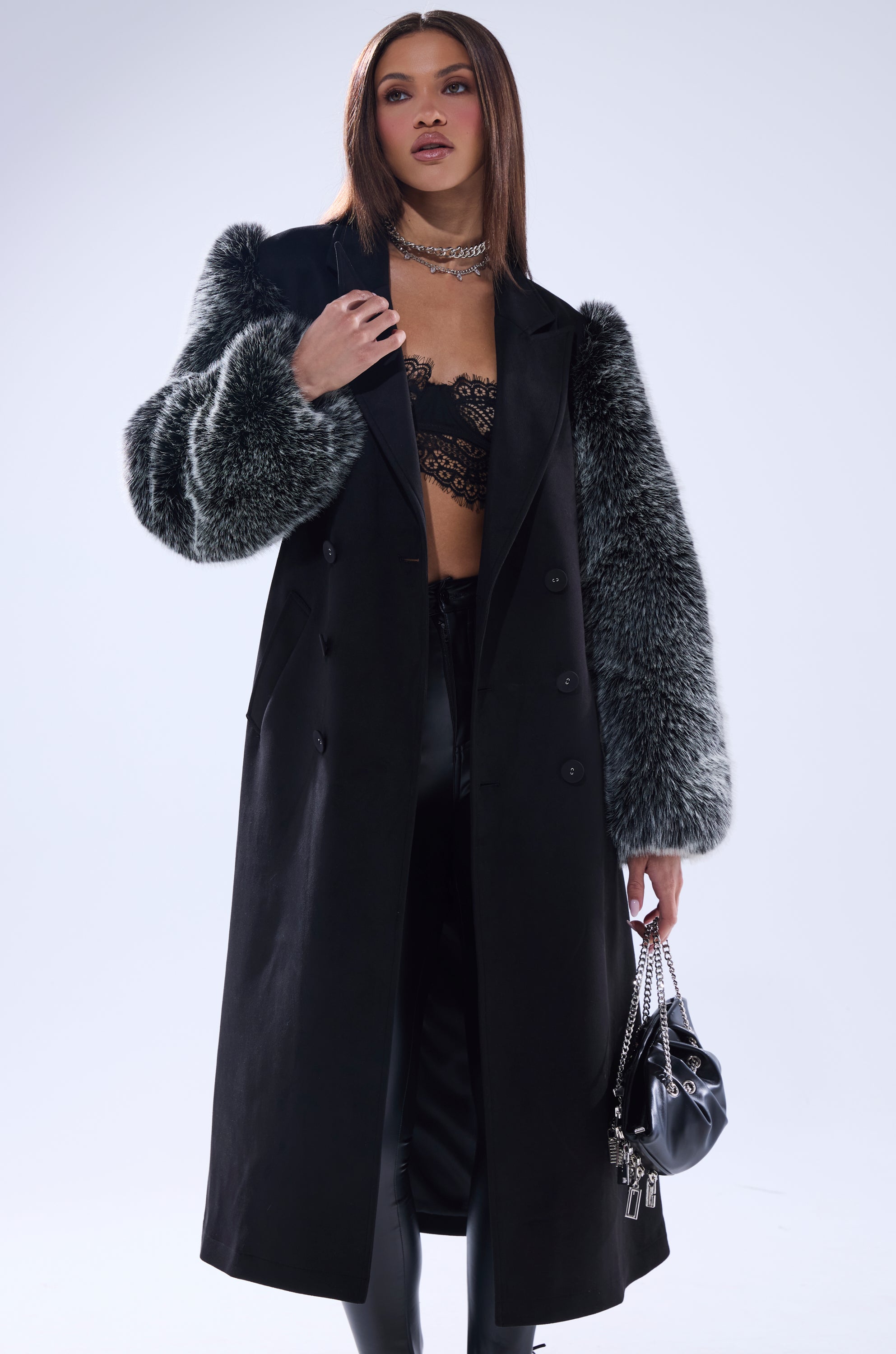 A woman in streetwear couture wears the NOLITA FUR SLEEVE TRENCH, a long black coat with gray fur sleeves, layered over a black lace bralette and pants. She carries a chain-strap bag and accessorizes with a silver choker.