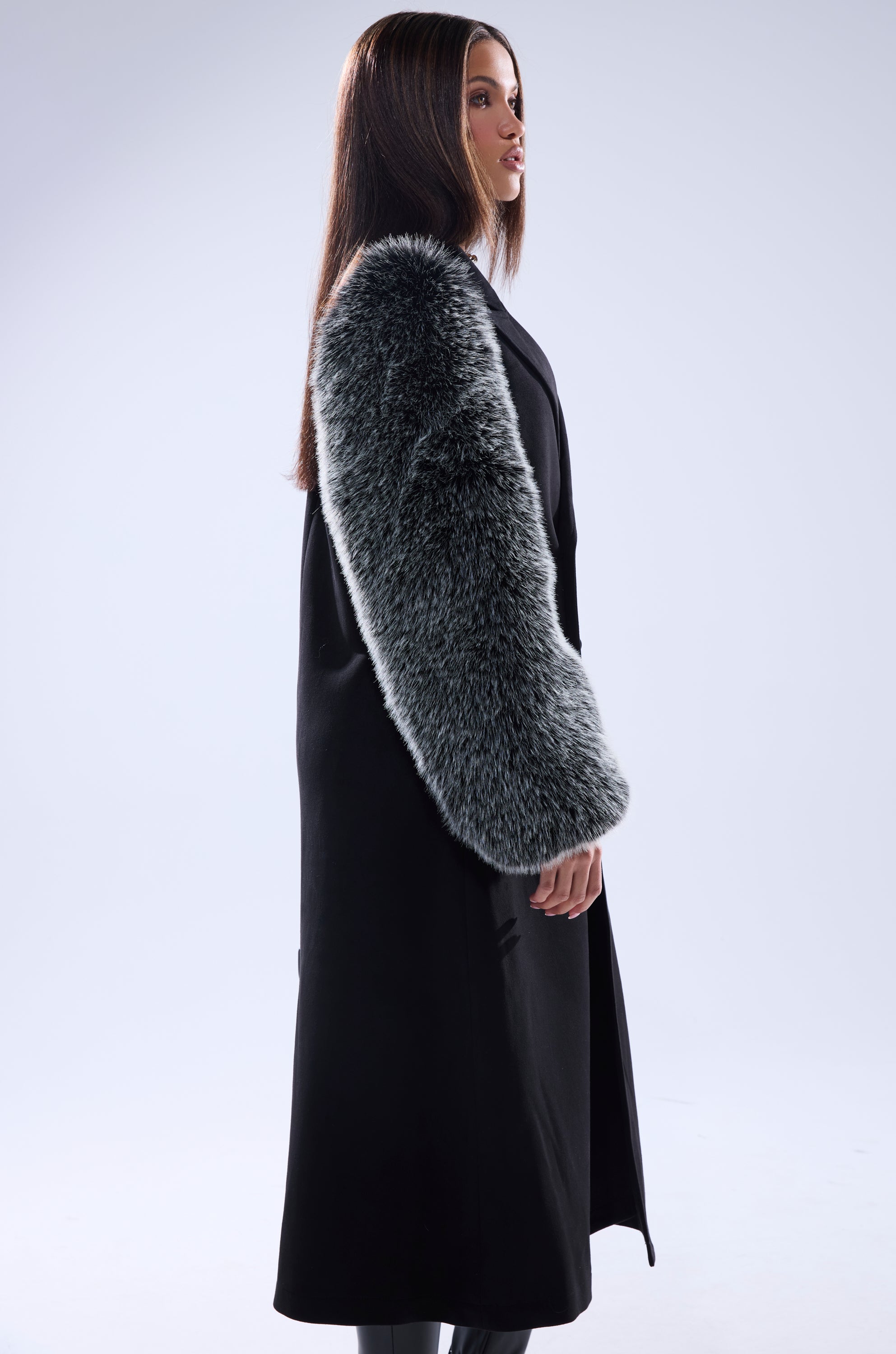 A woman in profile, with loose straight brown hair and a calm expression, wears the NOLITA FUR SLEEVE TRENCH—featuring large, fluffy gray sleeves and a longline silhouette—against a plain light background.