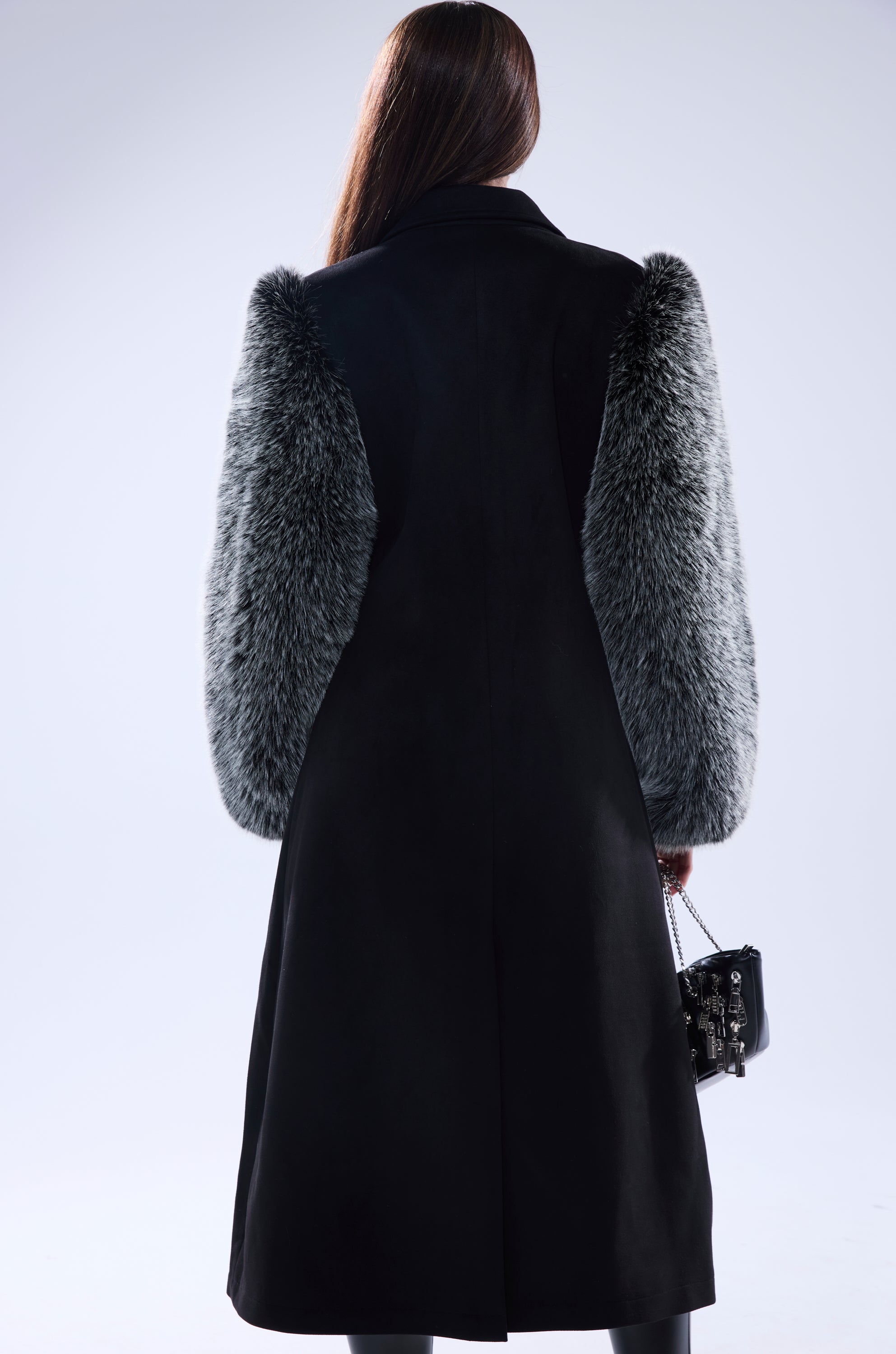 Seen from behind, a person with long brown hair wears the NOLITA FUR SLEEVE TRENCH—a faux fur trench coat with gray furry sleeves—holding a small black handbag. The striking longline silhouette pops against a plain, light backdrop.