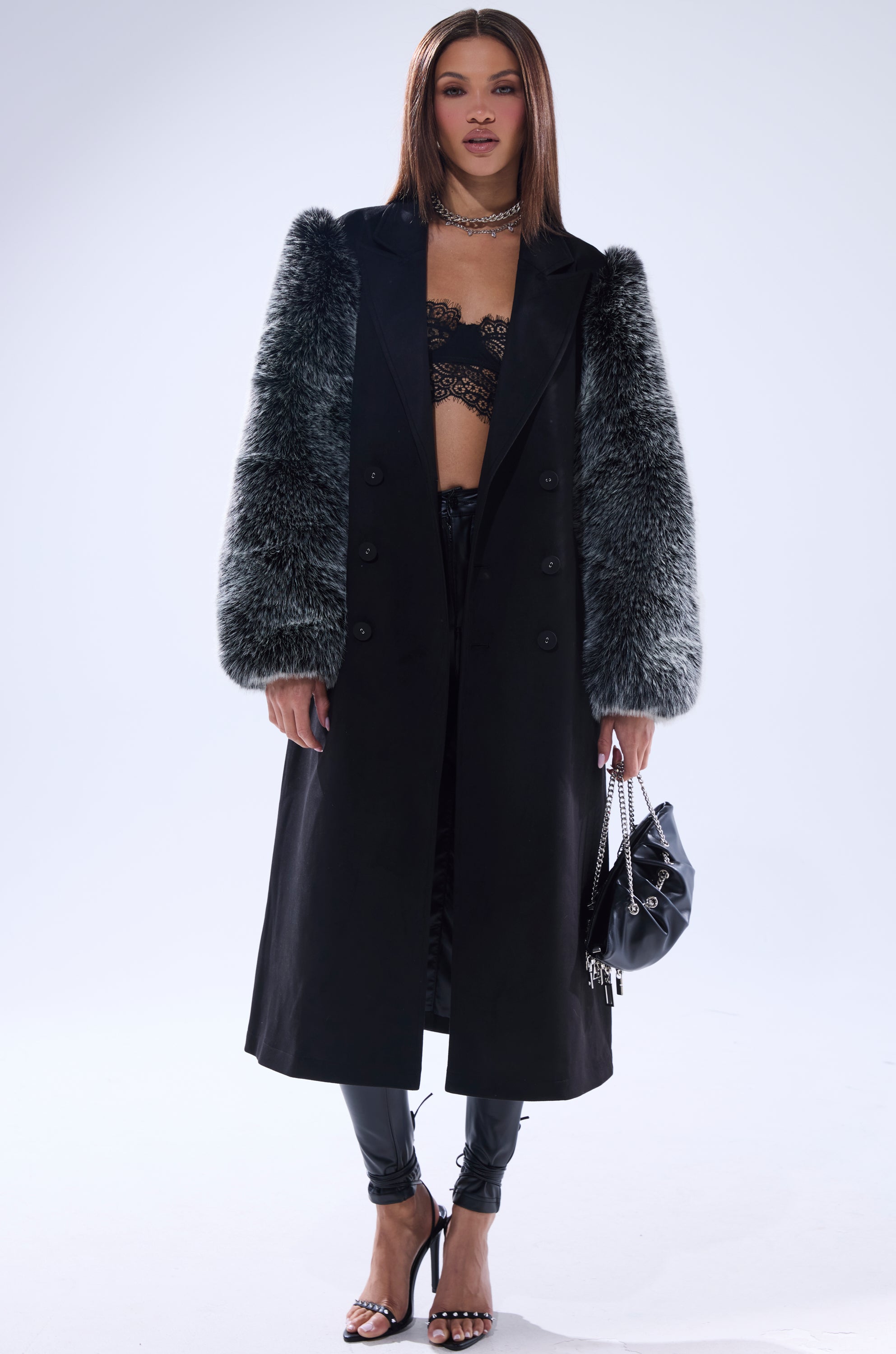 A woman models the NOLITA FUR SLEEVE TRENCH with textured gray sleeves, paired with a black lace bralette, black leather pants, heeled sandals, and a black handbag against a light background.