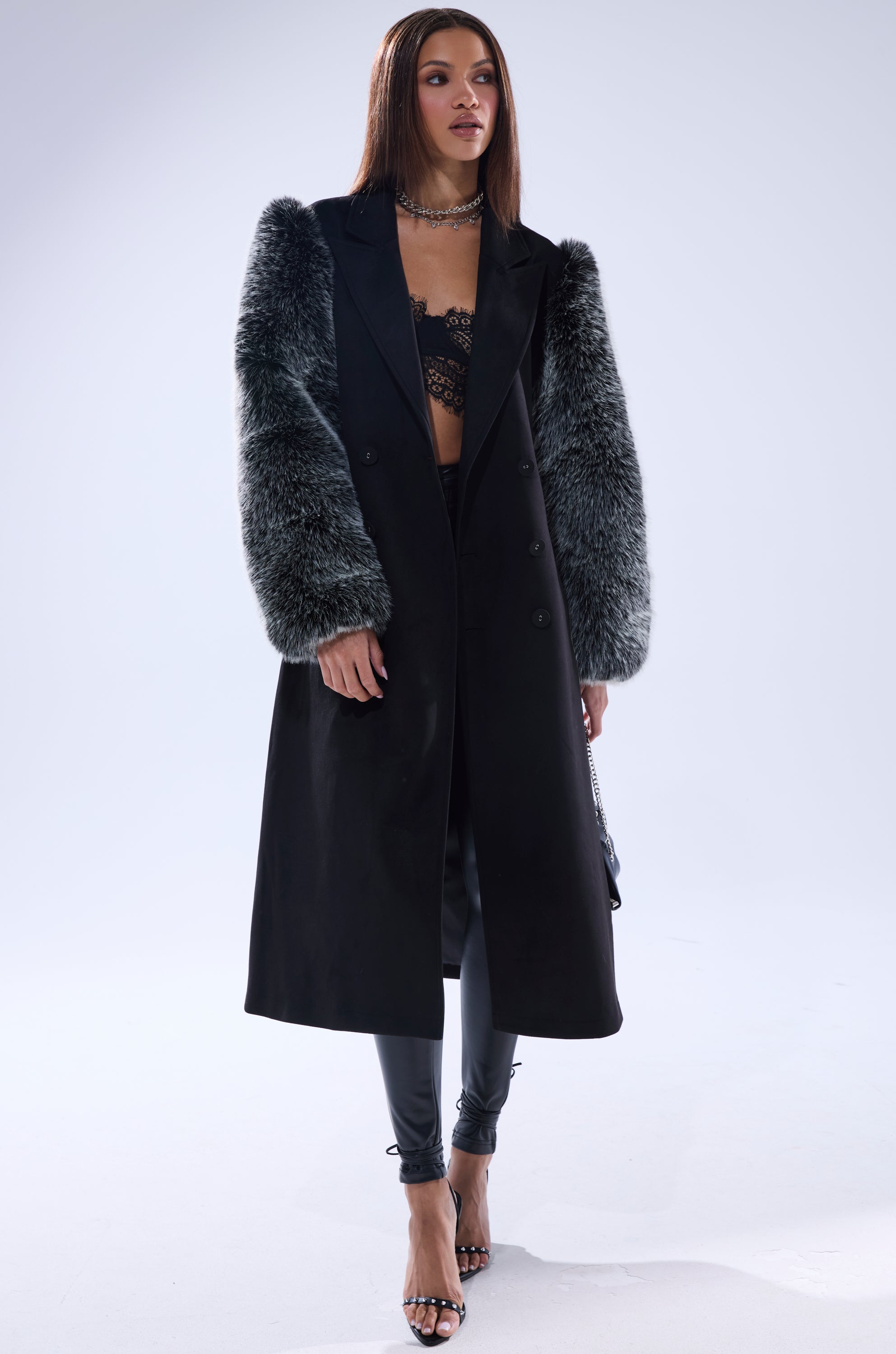 A woman models the NOLITA FUR SLEEVE TRENCH with gray fur sleeves, paired with a black lace bralette, black pants, and high-heeled sandals against a plain white background.