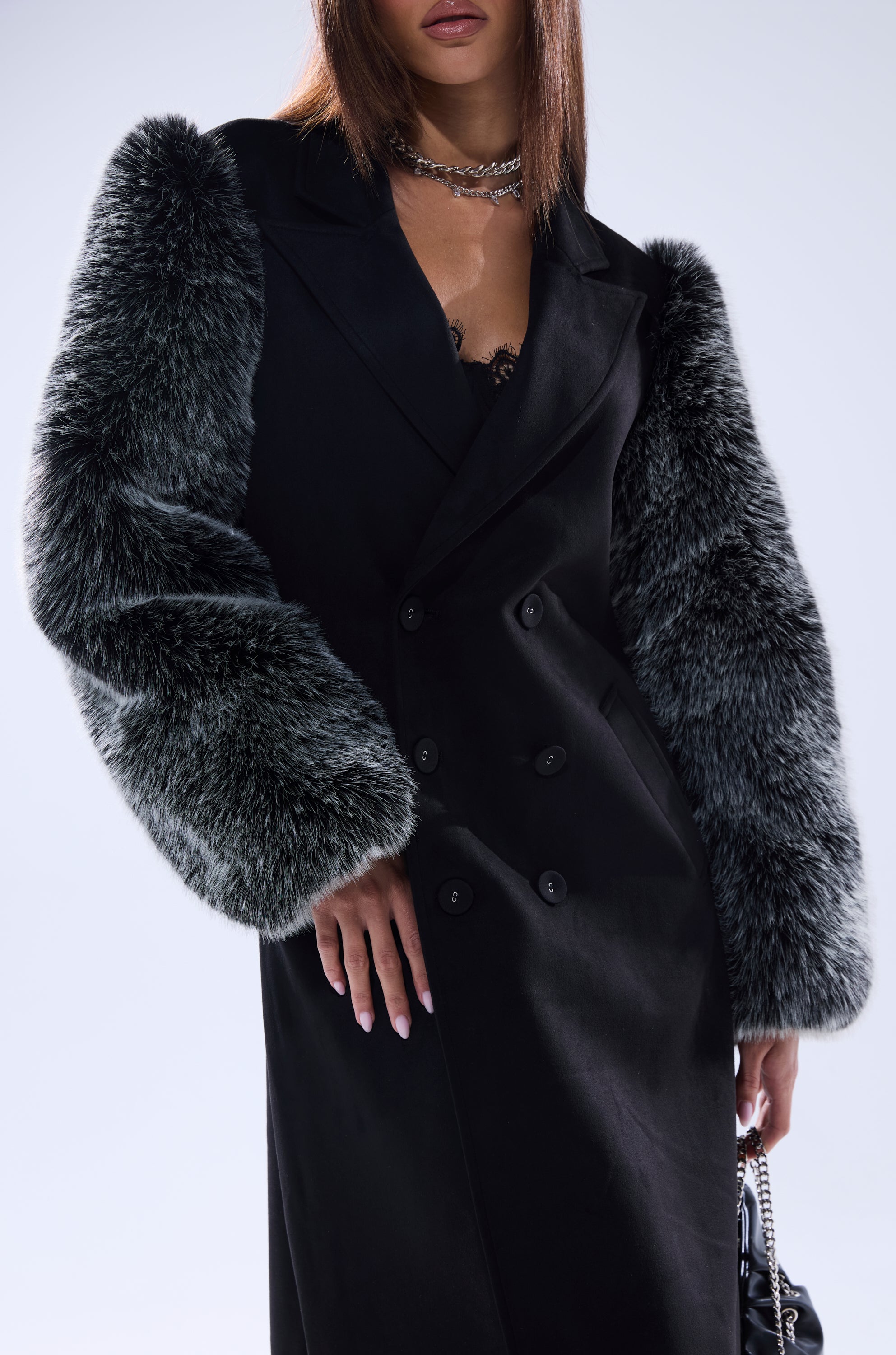 A woman with straight brown hair wears the NOLITA FUR SLEEVE TRENCH, a black trench coat with textured gray faux fur sleeves and a longline fit. She accessorizes with layered necklaces and a black handbag; image cropped mid-face down.