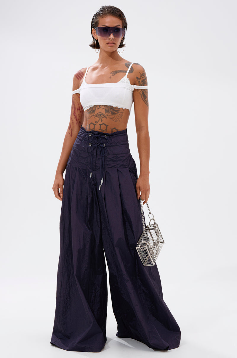 A tattooed individual in a white crop top, DANCE THE NIGHT AWAY DENIM PALAZZO PANT with a wide-leg fit, large sunglasses, and holding a small geometric metallic handbag poses against a plain light background.