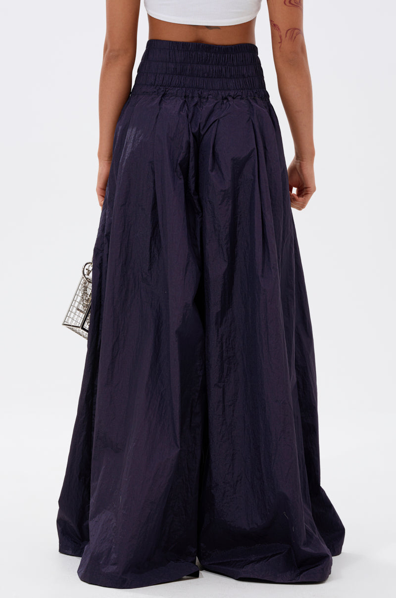 A person wears the DANCE THE NIGHT AWAY DENIM PALAZZO PANT in dark navy, featuring a high-rise fit, wide-leg silhouette, and gathered elastic waistband. Seen from the back with a small handbag and a white crop top.